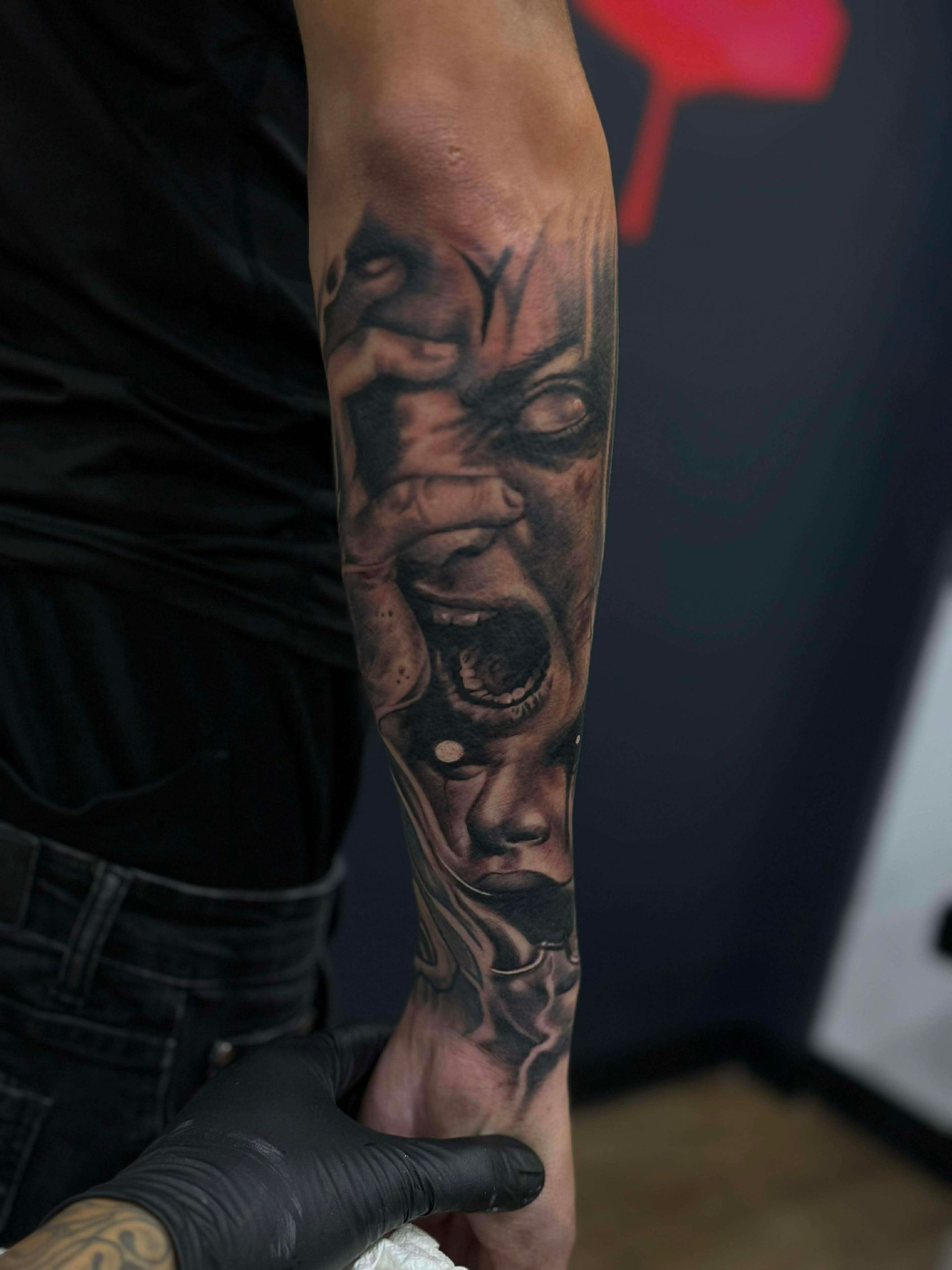 Tattoo of a distorted face in grayscale on forearm. A hand wearing a black glove holds the wrist. Dark background.