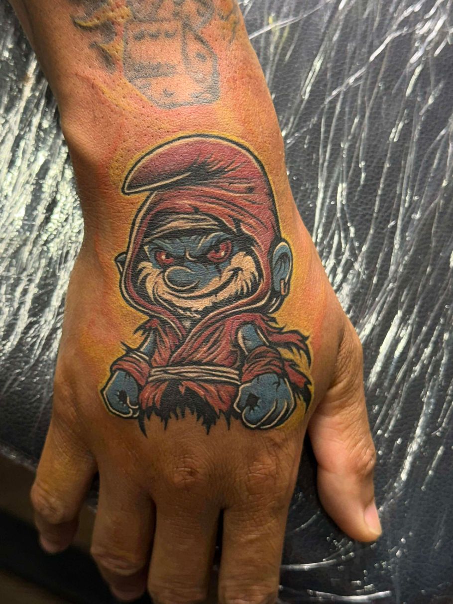 Tattoo of a menacing Smurf wearing a red hooded robe on a hand.