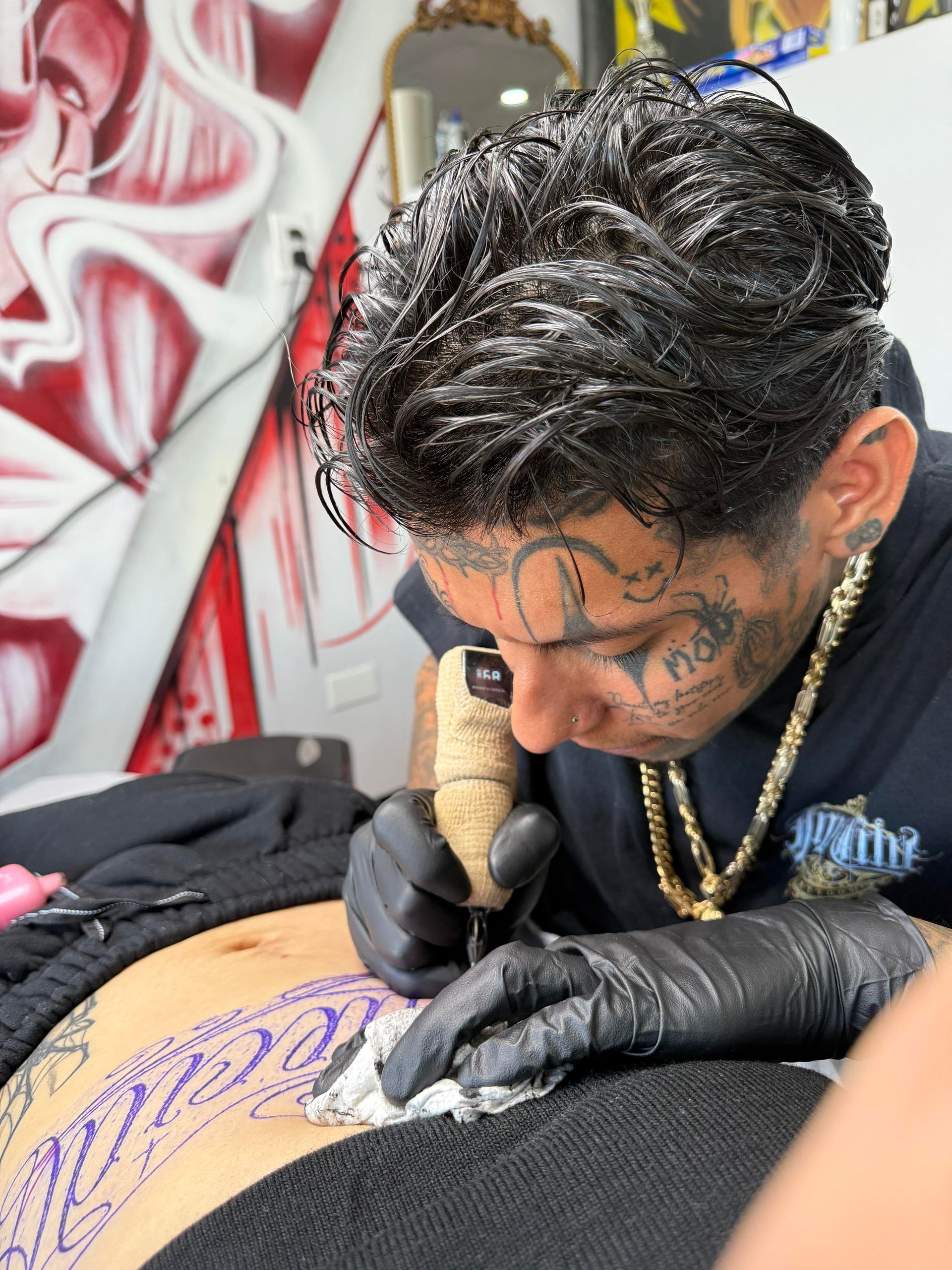 Tattoo artist with face tattoos, inking a design on a person's abdomen, in a tattoo studio.