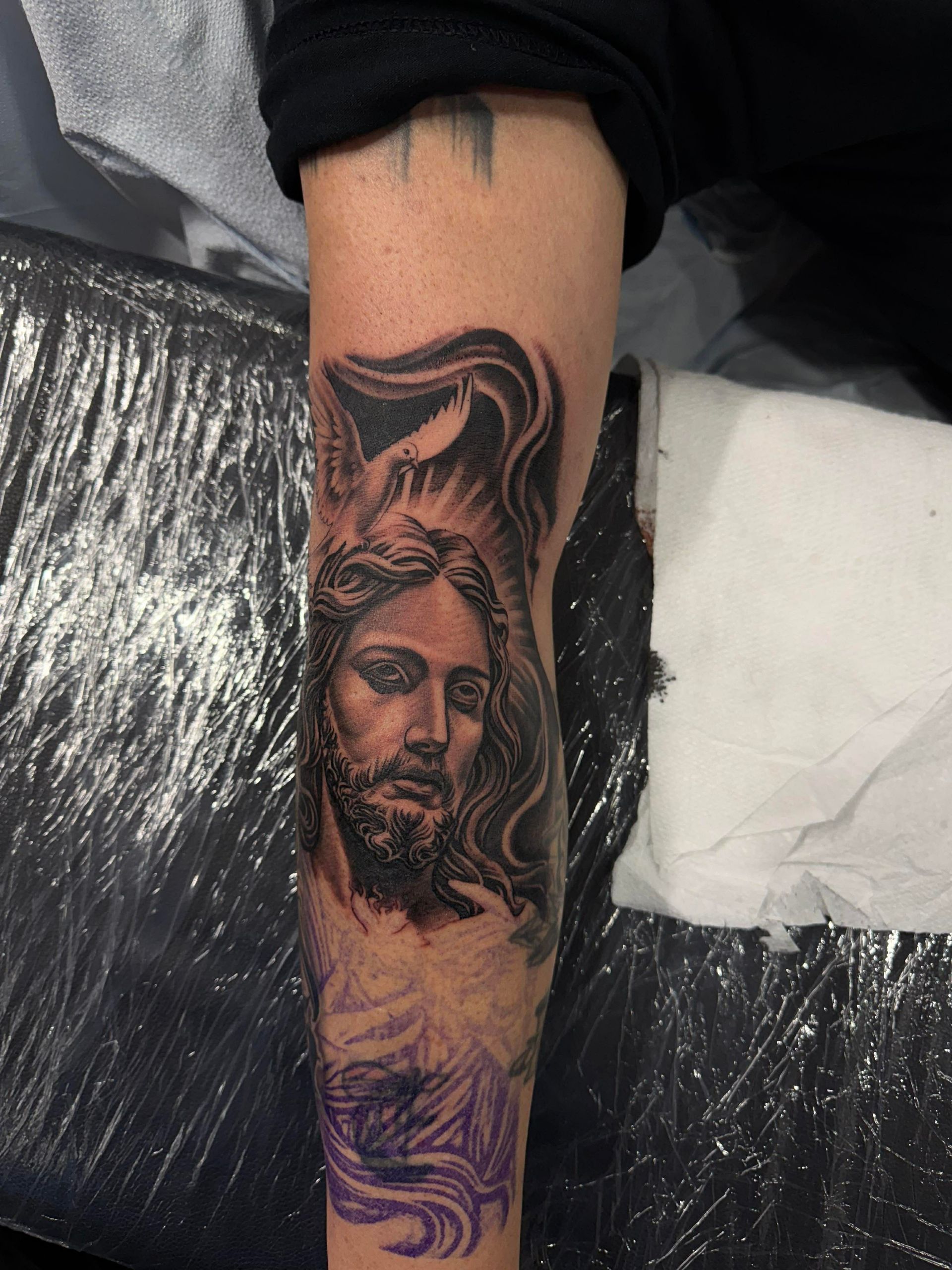 Tattoo of Jesus on a leg. Black and gray portrait with flowing hair, a beard, and a halo.