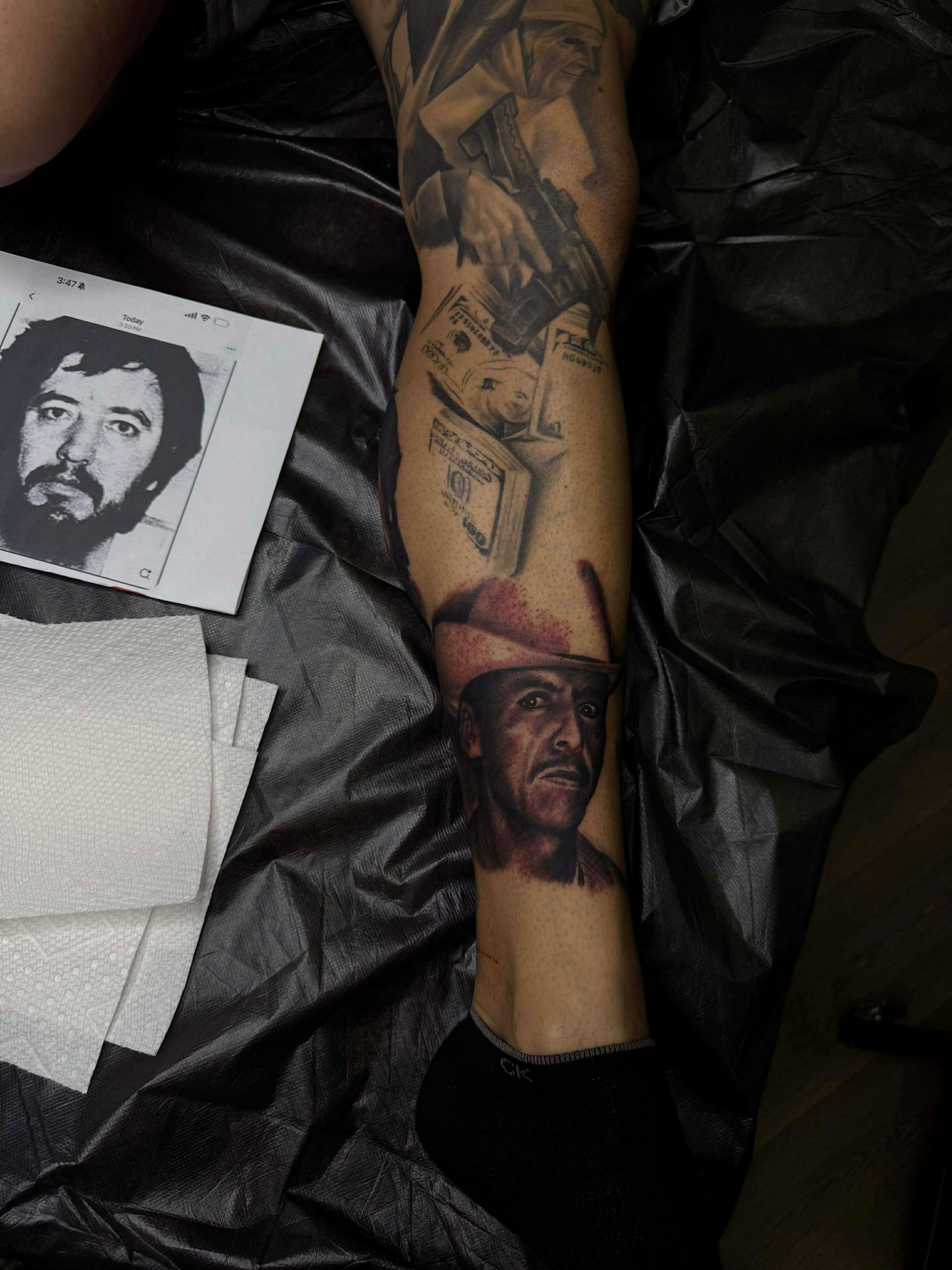 Tattoo of a man's face on leg. Reference photo beside. Other tattoos and cash also visible.