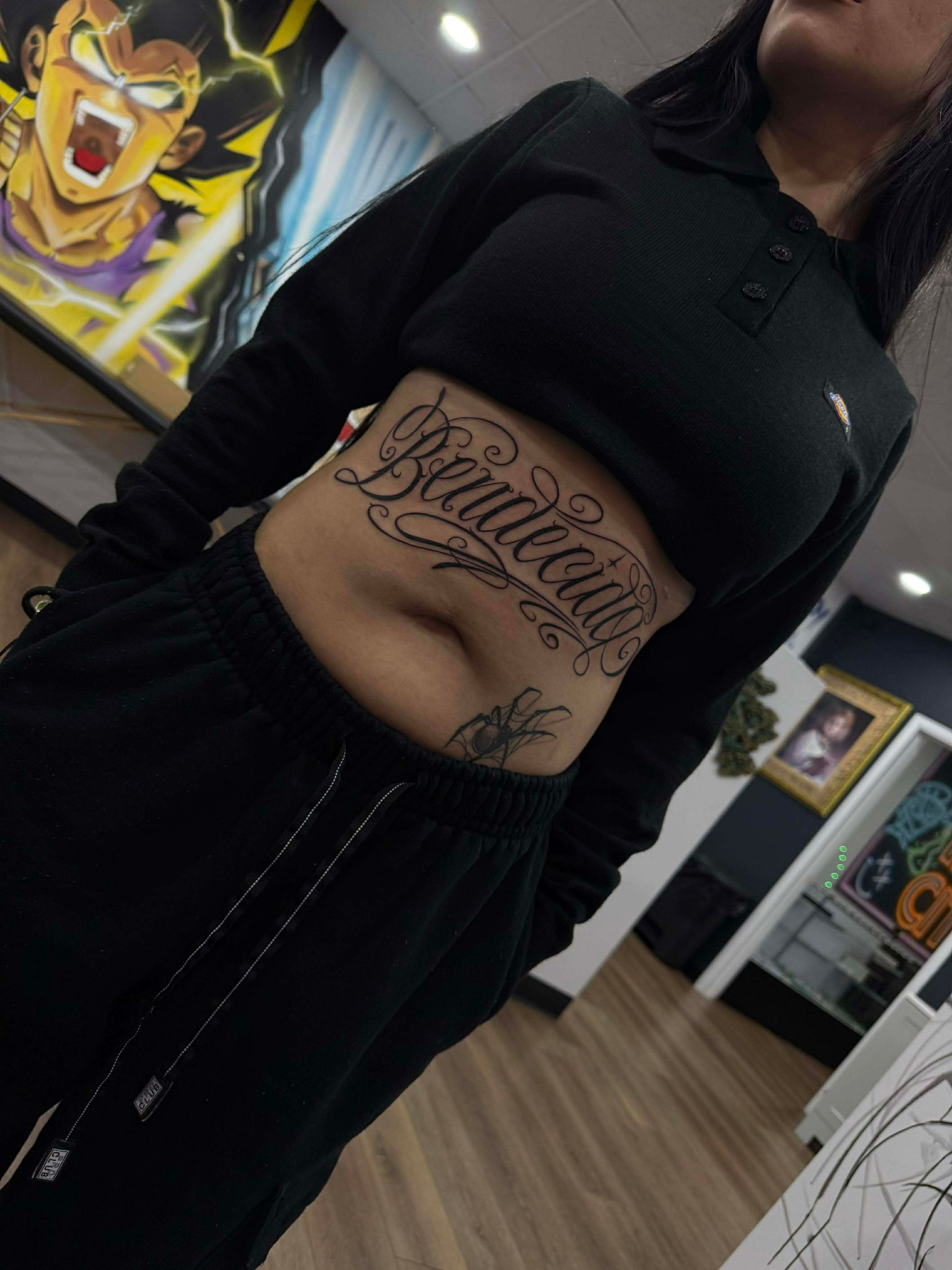 Woman with a stomach tattoo, wearing a black crop top and sweatpants, standing in a tattoo parlor.