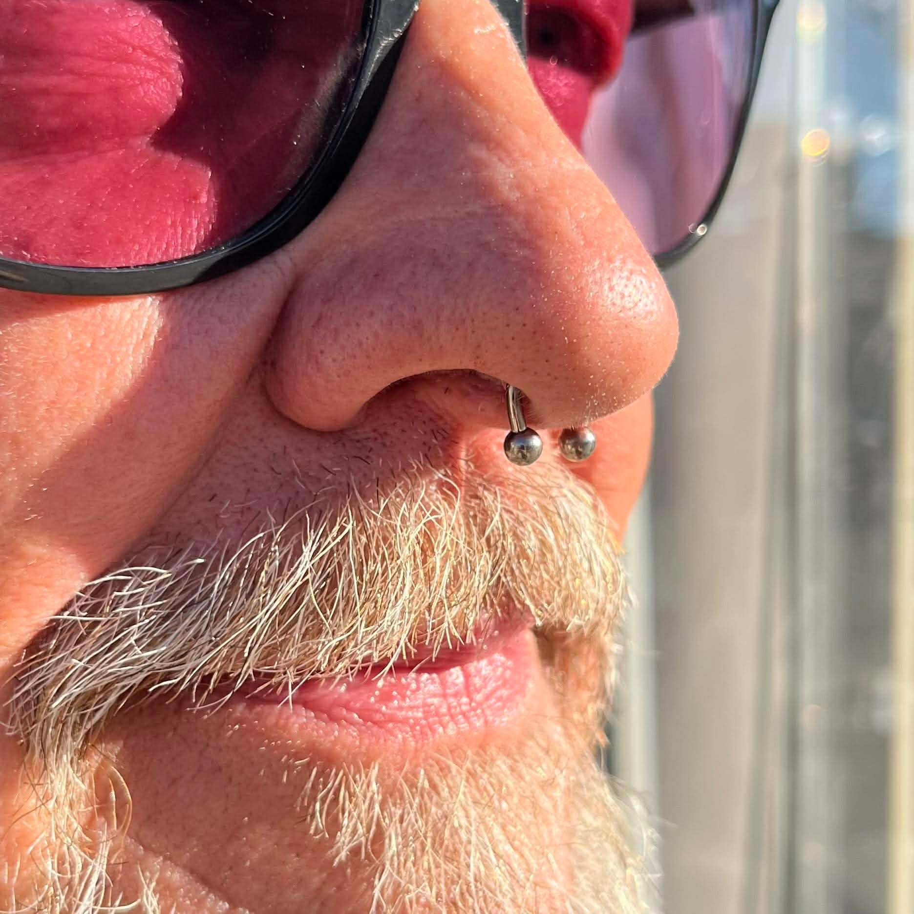 Close-up of a person's face wearing sunglasses, a nose ring, and a light-colored mustache.