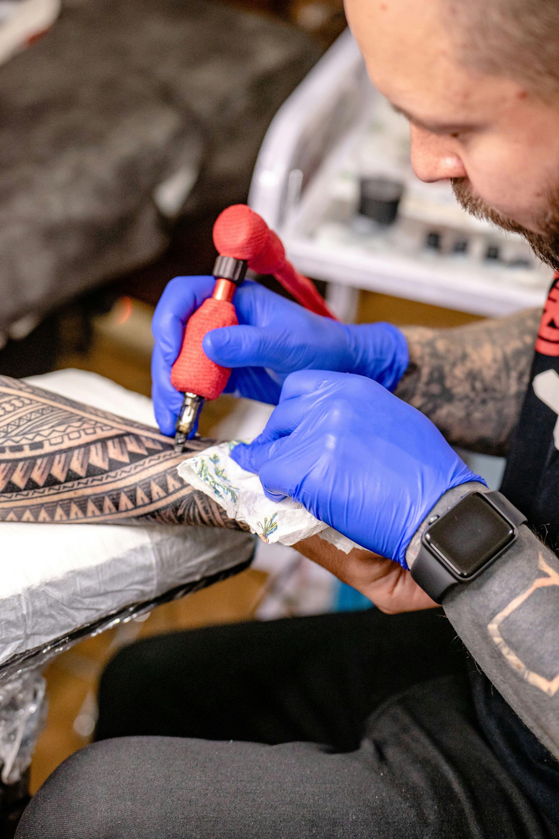 Tattoo artist in blue gloves, using a tattoo machine on a person's arm, inside a studio.