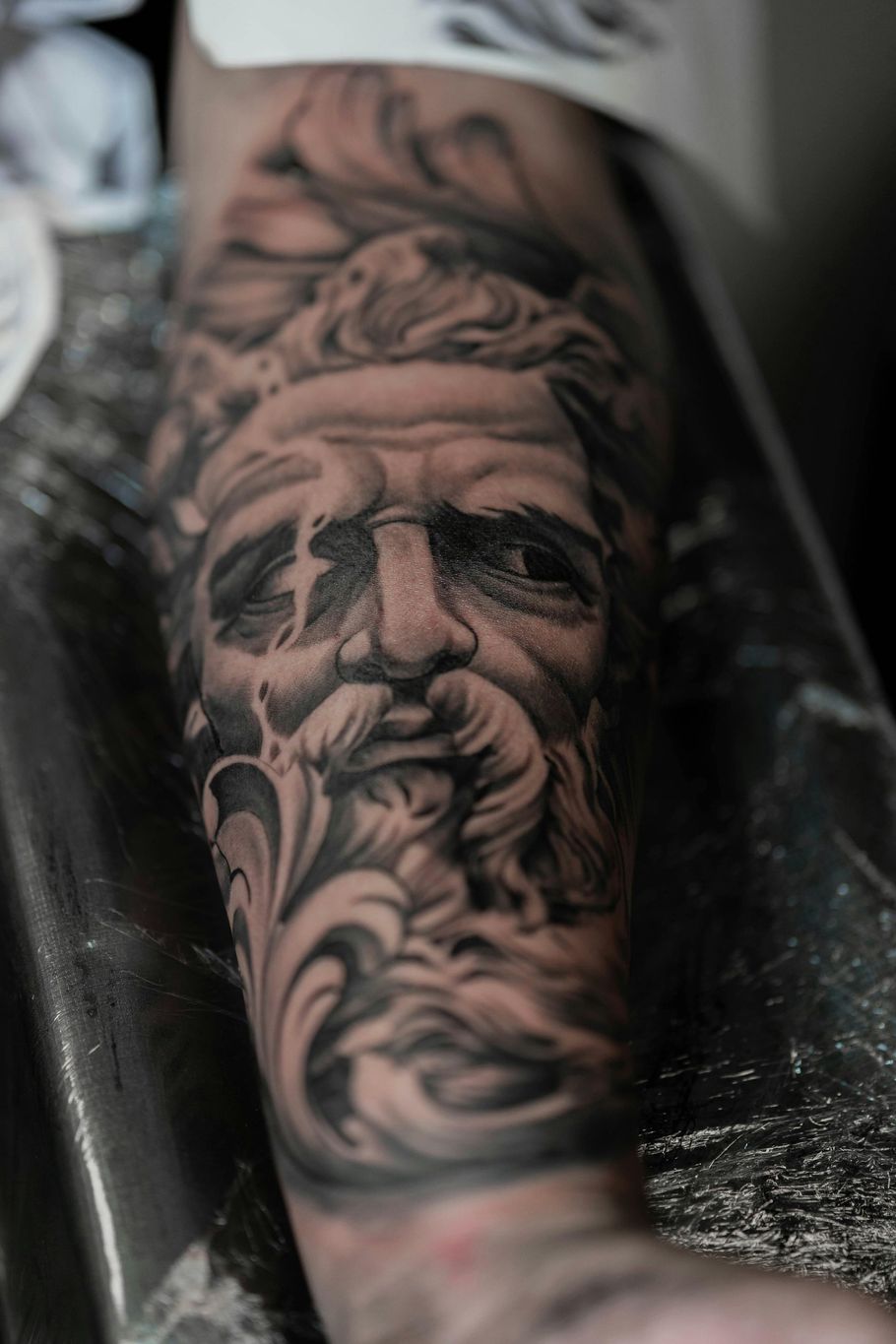 Close-up of a detailed black and grey tattoo of a bearded face with elaborate swirling detail.
