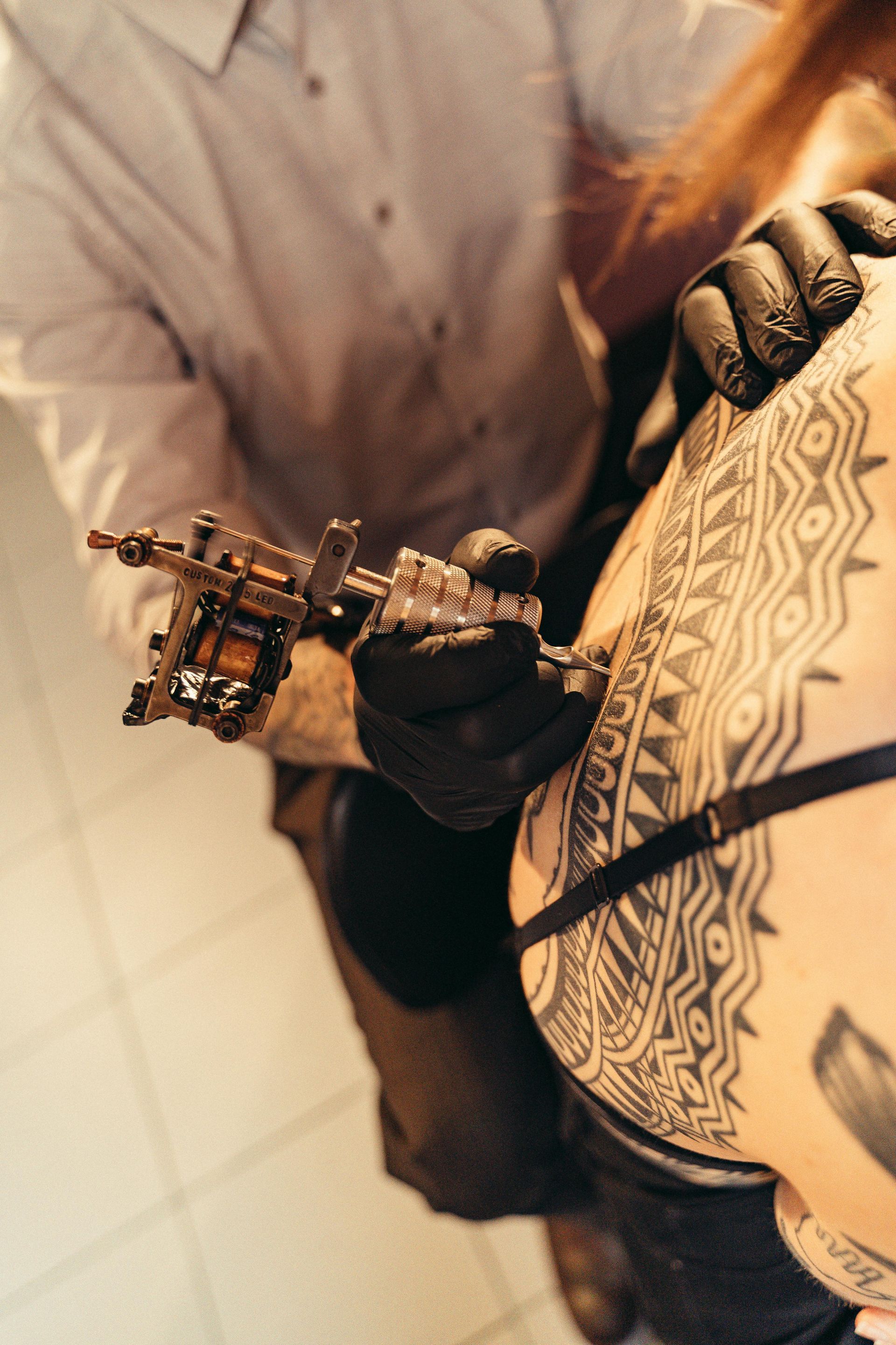 Tattoo artist in black gloves, using a tattoo machine on a person's upper back with tribal patterns.