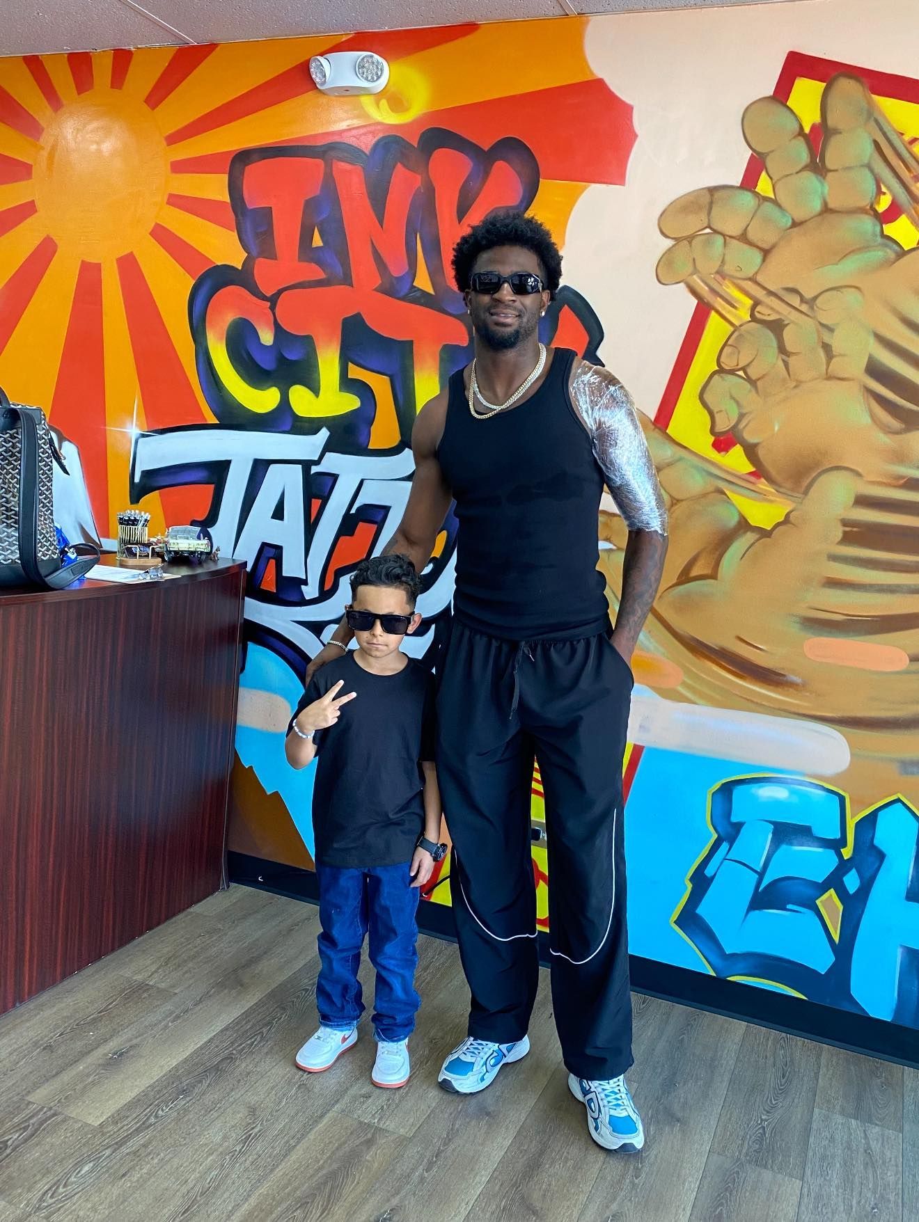 Man and child pose in tattoo shop, with graffiti art wall behind them. Both wear black, child gives peace sign.