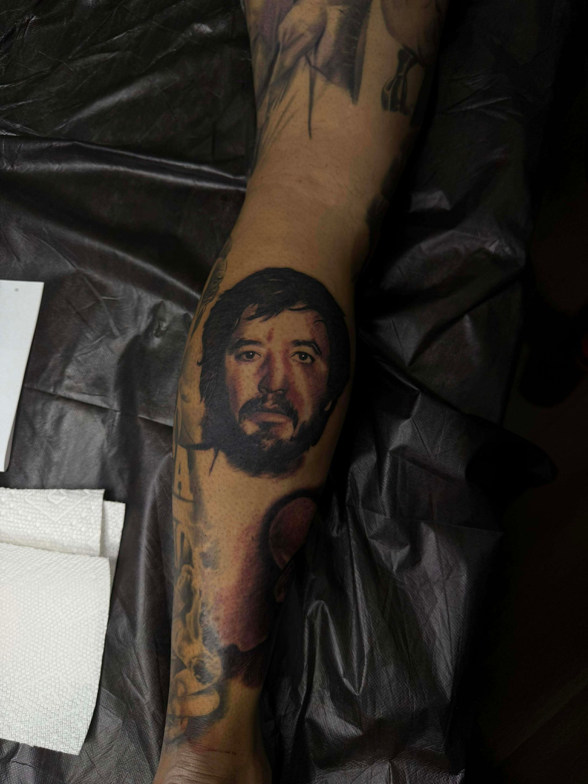 Tattoo of a man's face on an arm. The man has dark hair and a beard, with a neutral expression.