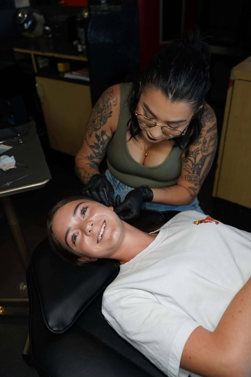 Woman getting an ear piercing from a person with black gloves and tattoos at a studio.