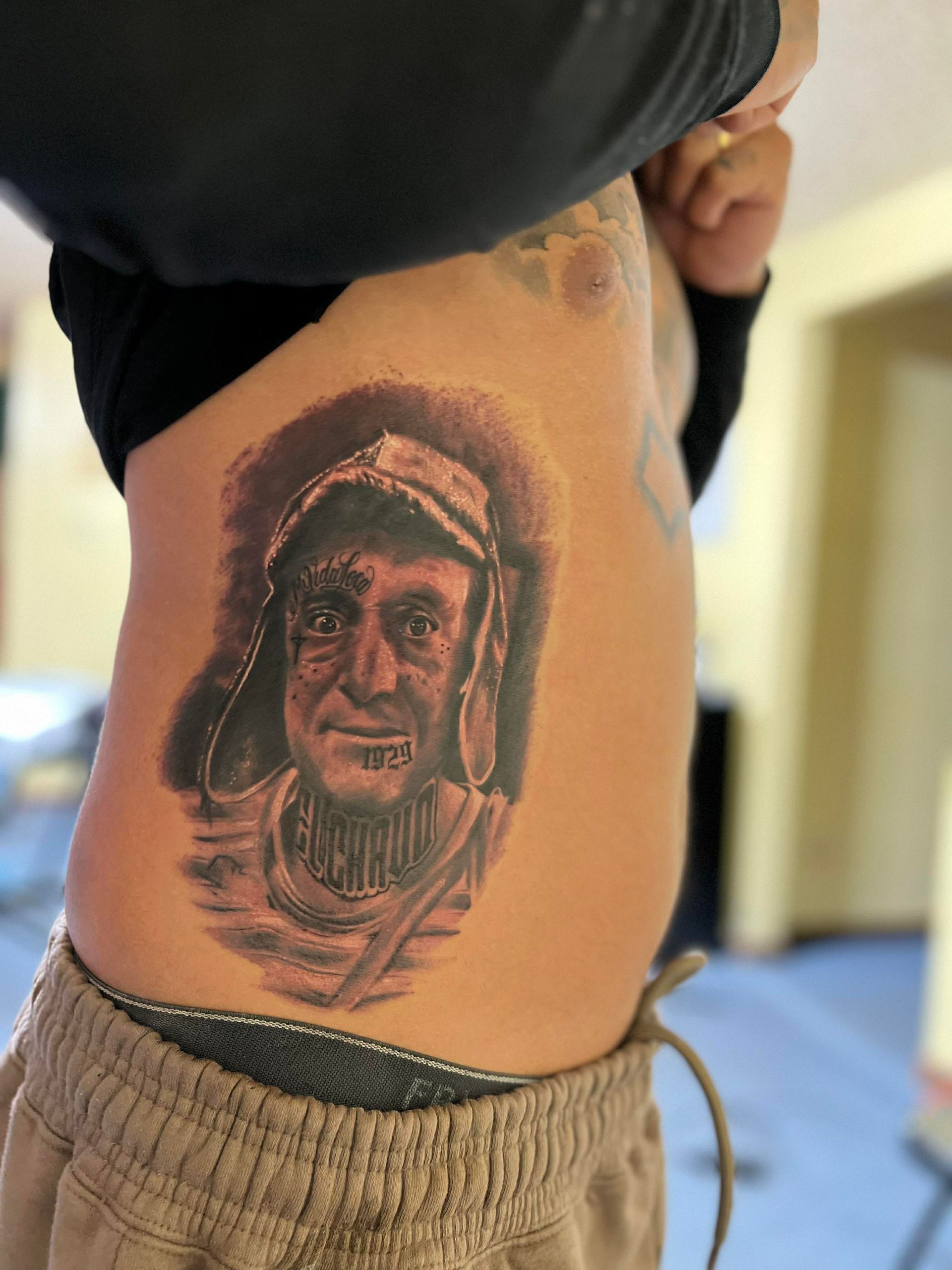 Tattoo of a man on a person's side, gray ink, wearing a hat, with a slightly smiling expression.