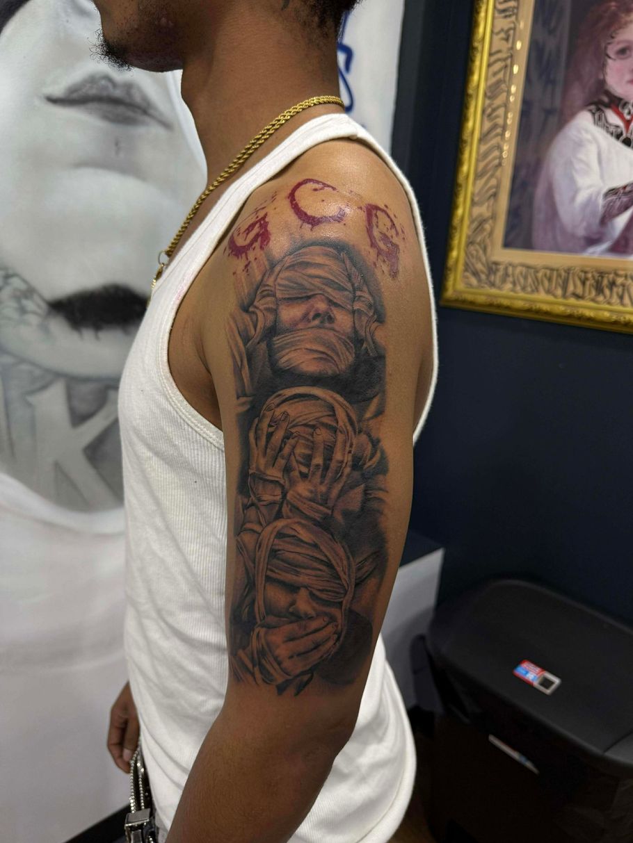 Man with a detailed tattoo of three figures covering eyes, ears, and mouth on arm. Gold chain. Tank top. Studio setting.