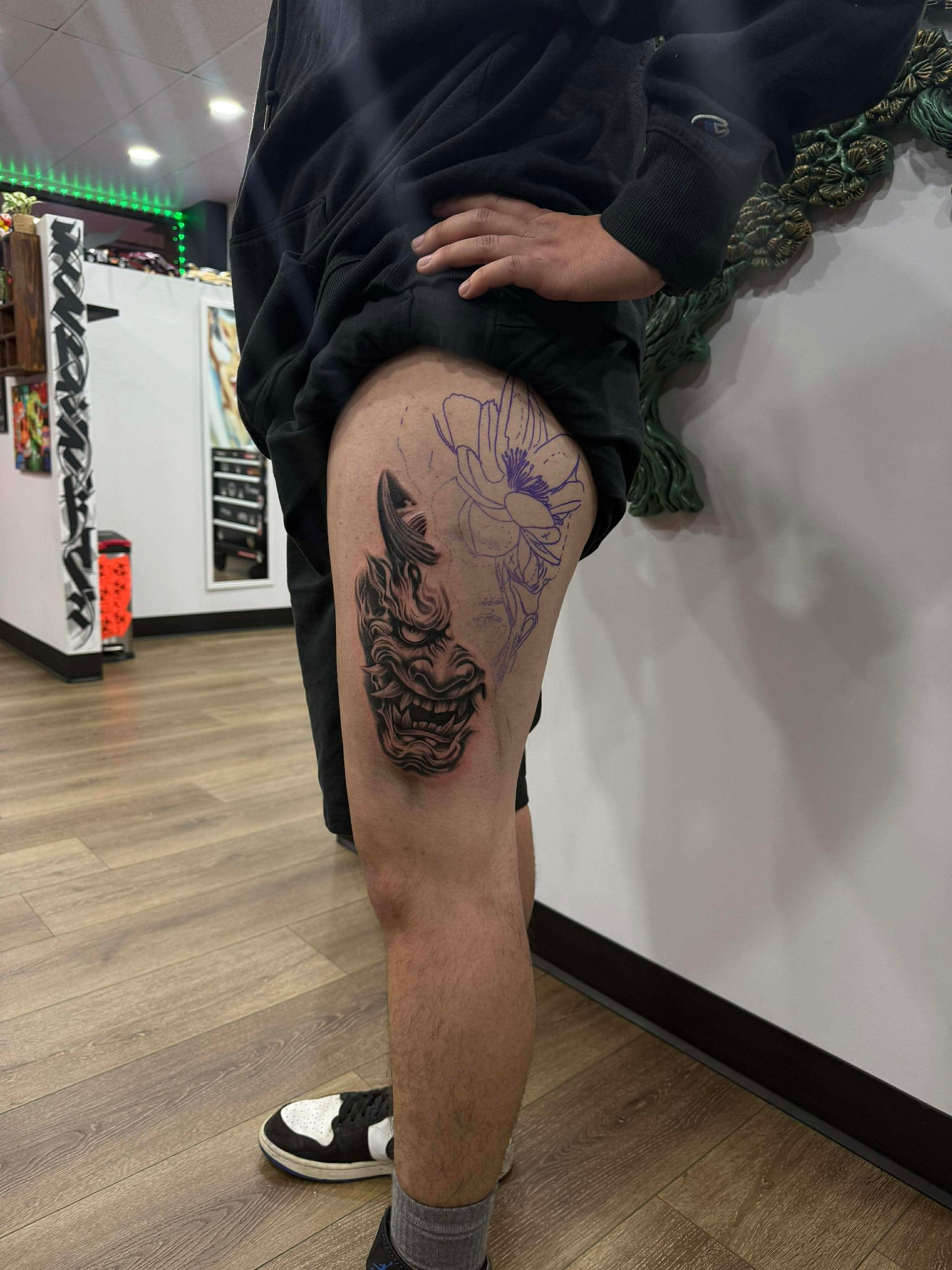 Person with a leg tattoo, dark, textured design. Standing indoors near a wall, wearing shorts and sneakers.