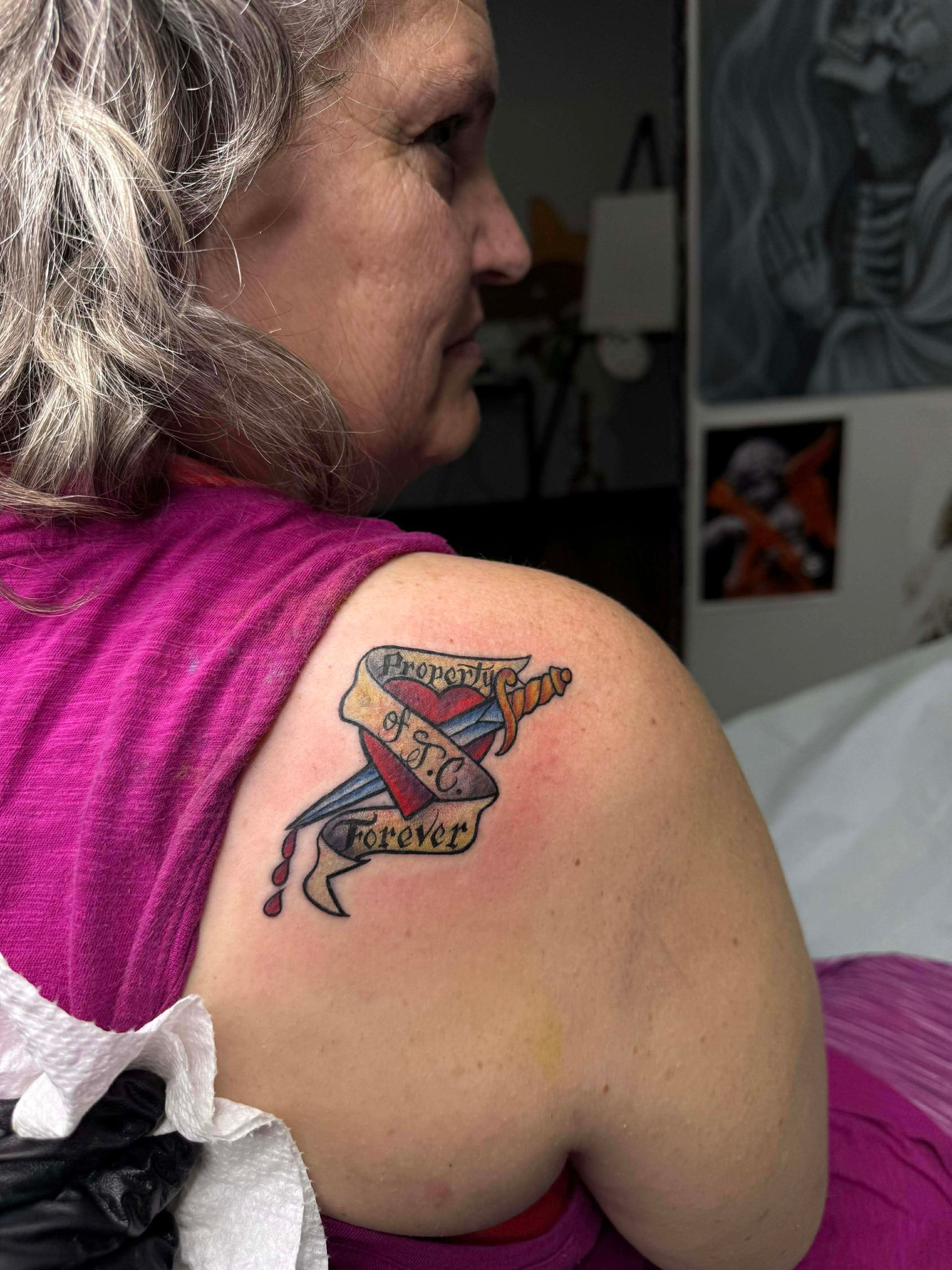 Woman with shoulder tattoo: heart pierced by dagger, wrapped in banner text.