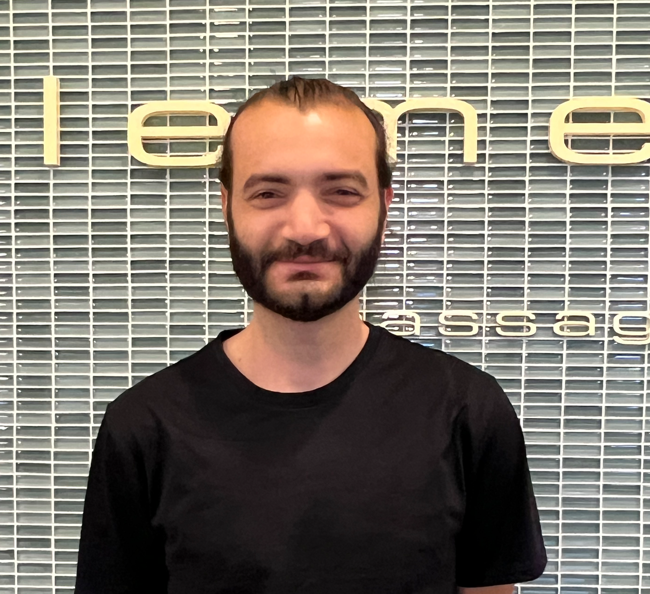 A man with a beard wearing a black shirt stands in front of a wall that says iteme