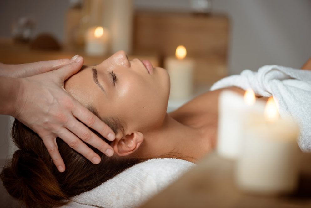 A woman is getting a head massage in a spa with candles in the background.