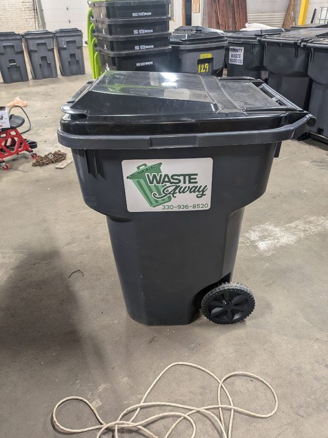 A black trash can with a sticker on it that says `` waste recycling '' is sitting on the ground.