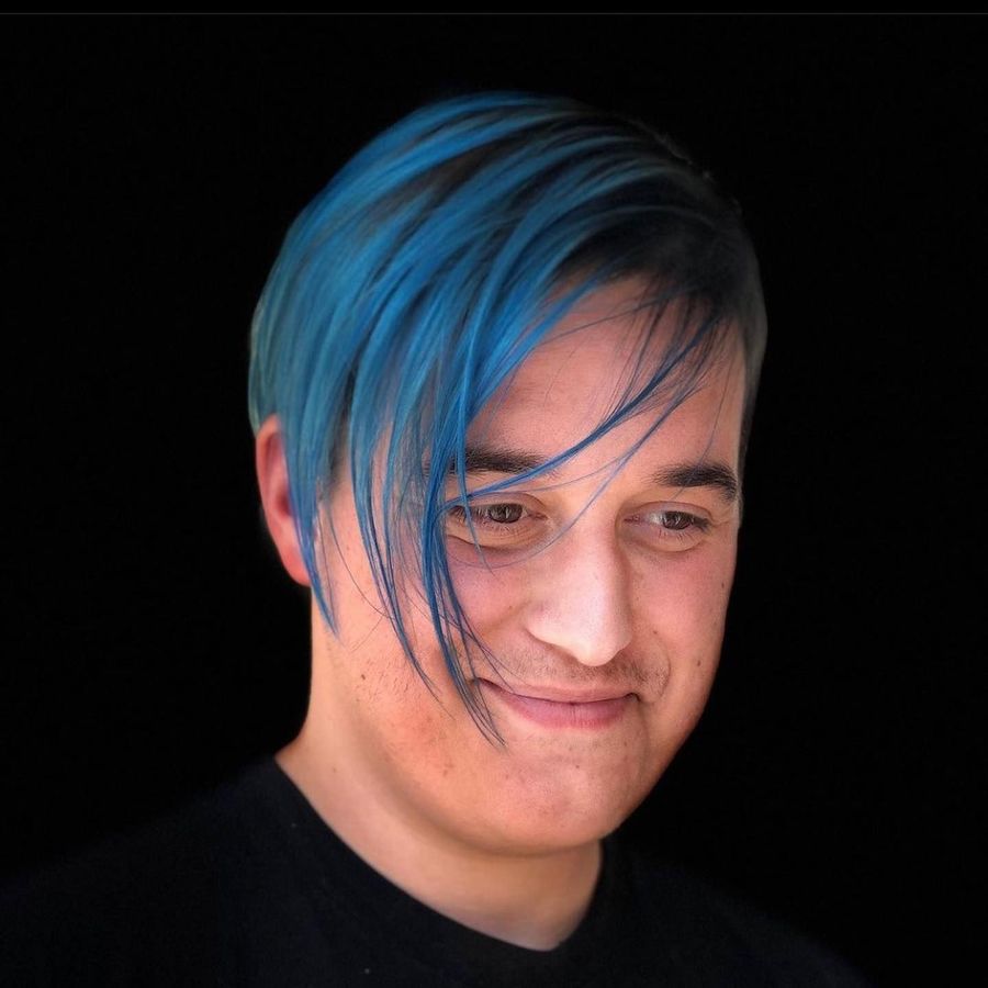 Man with blue hair, wearing a black shirt, looking down with a slight smile against a black background.