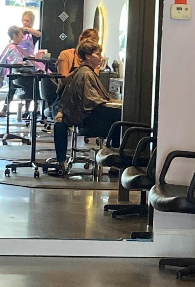 A woman in a salon getting her hair styled; other clients and stylists are visible in the background. The salon has a large mirror.