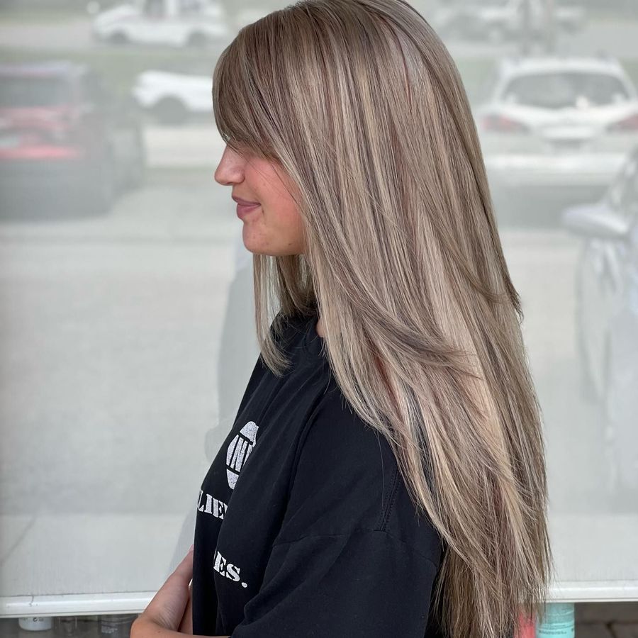 Woman with long, layered light brown hair and bangs, smiling. She wears a black t-shirt. Outside, with cars visible in the background.