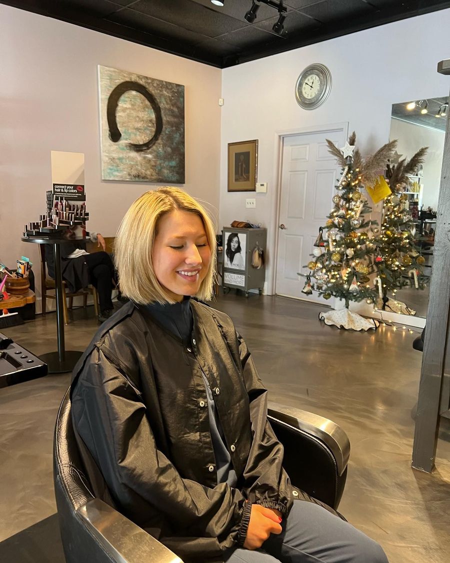 A young woman with blonde hair smiles while sitting in a salon chair, wearing a black cape. A Christmas tree decorates the background.