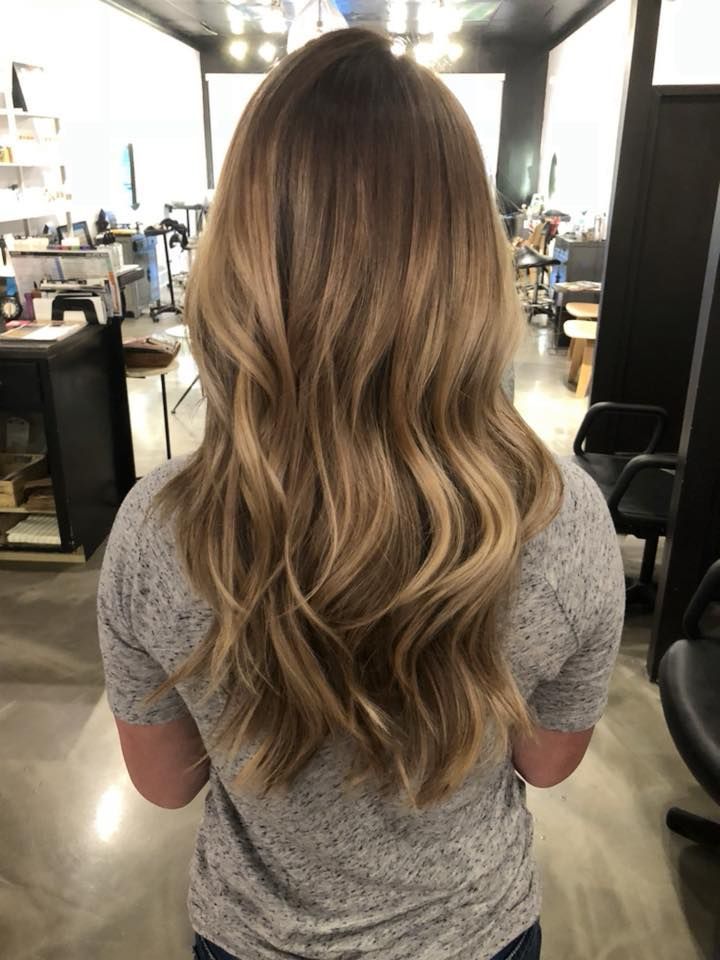 Woman with long, wavy blonde and brown hair, standing in a salon, wearing a gray t-shirt.