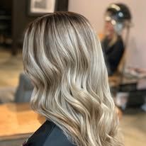 Blonde hair with brown highlights, styled in loose waves. The person is facing away, seated in a salon.