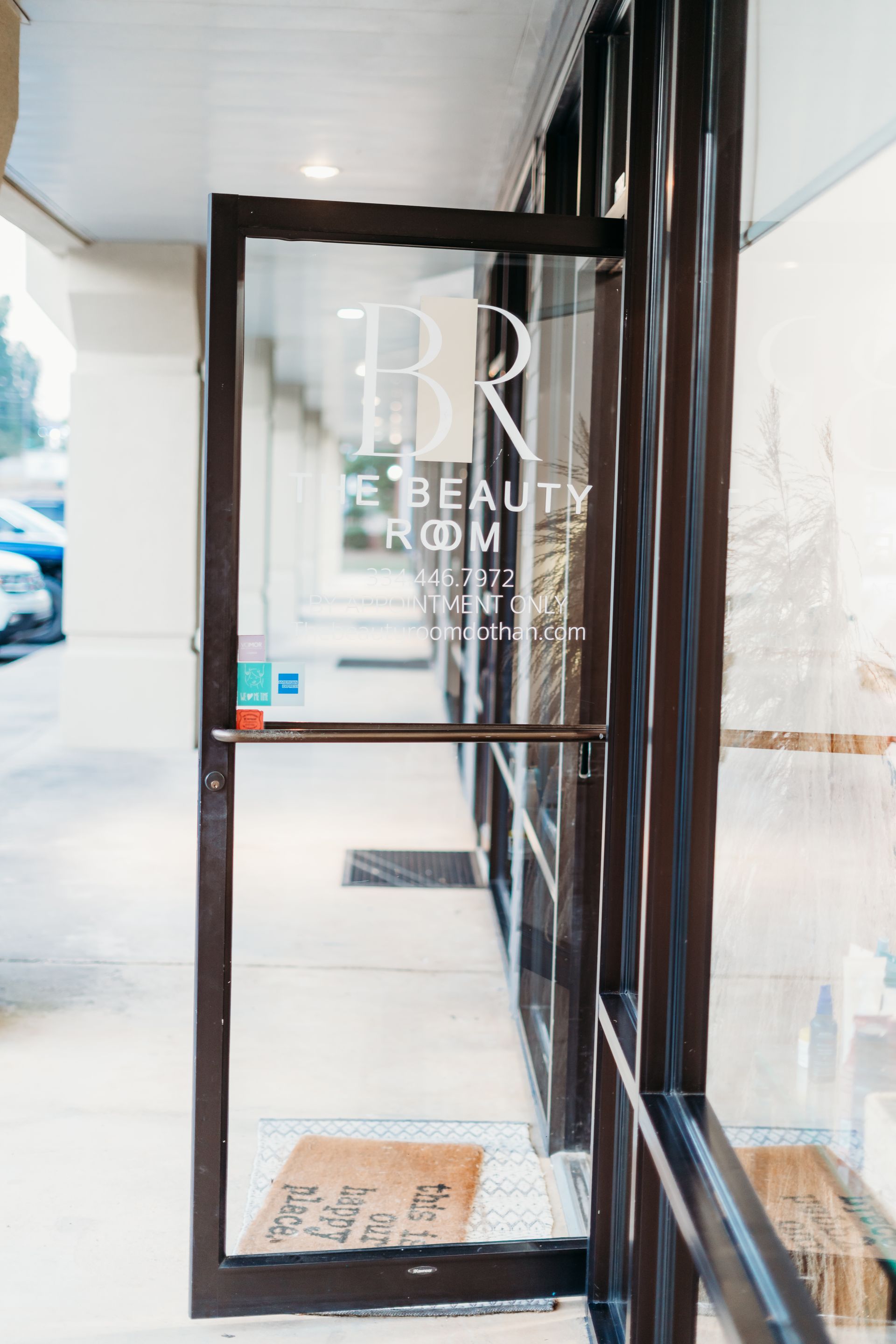 Glass door to a beauty room with logo, partially open. Brown frame, welcome mat, and a glimpse of the interior.