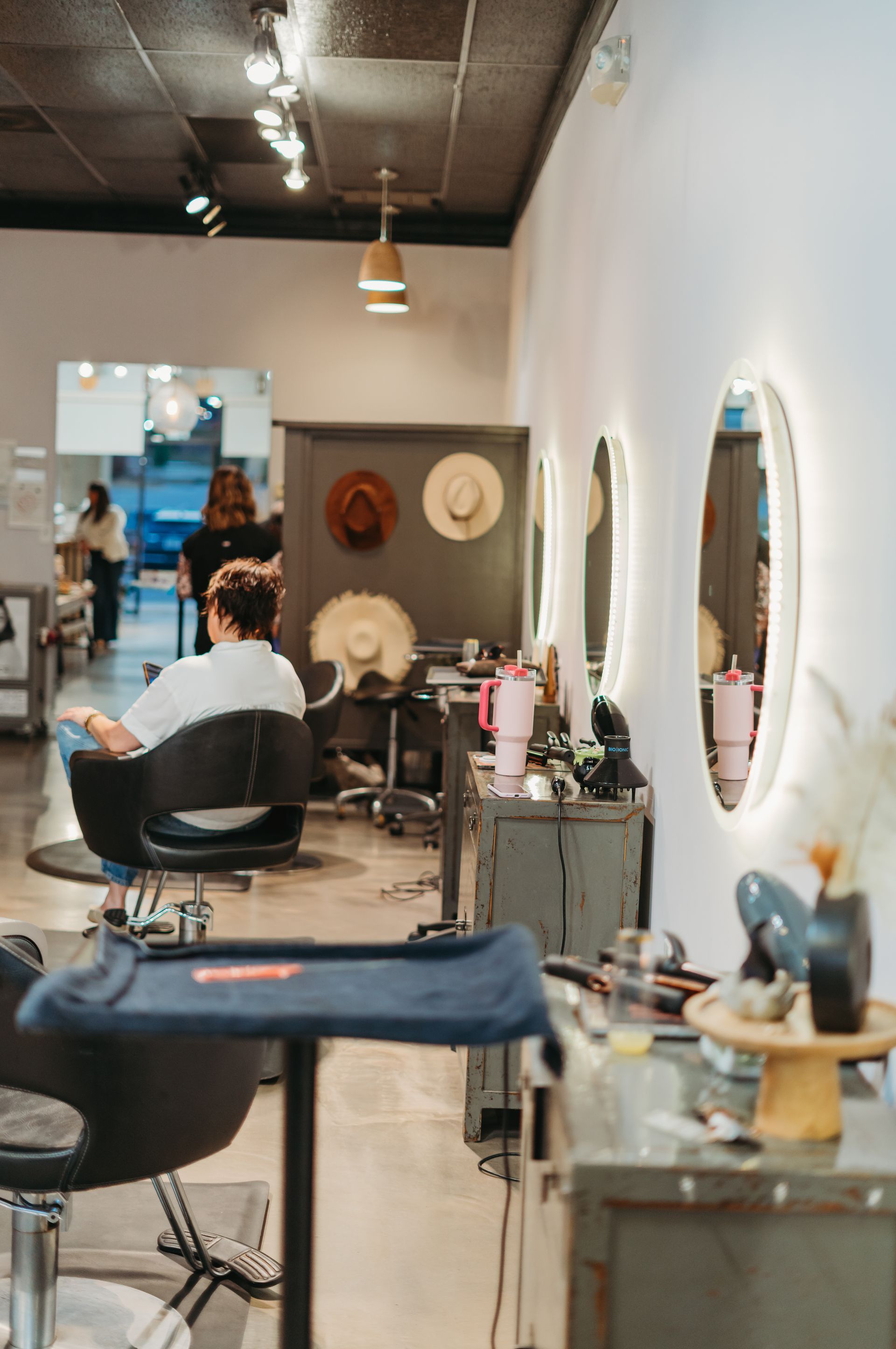 Hair salon interior.  Client in chair, stylist working.  Oval mirrors, hats on wall, and styling stations.