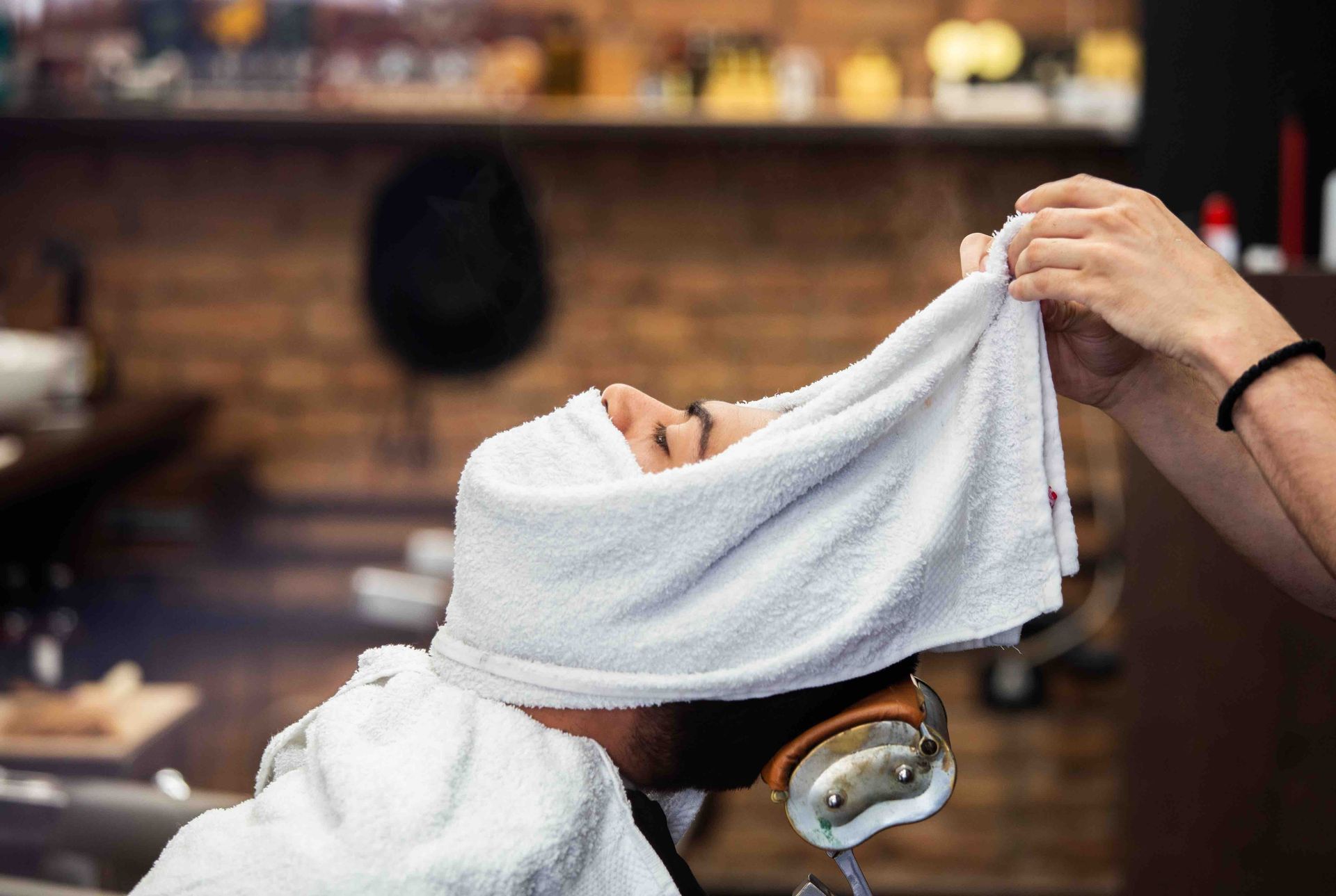A man is getting a shave at a barber shop with a towel wrapped around his head.
