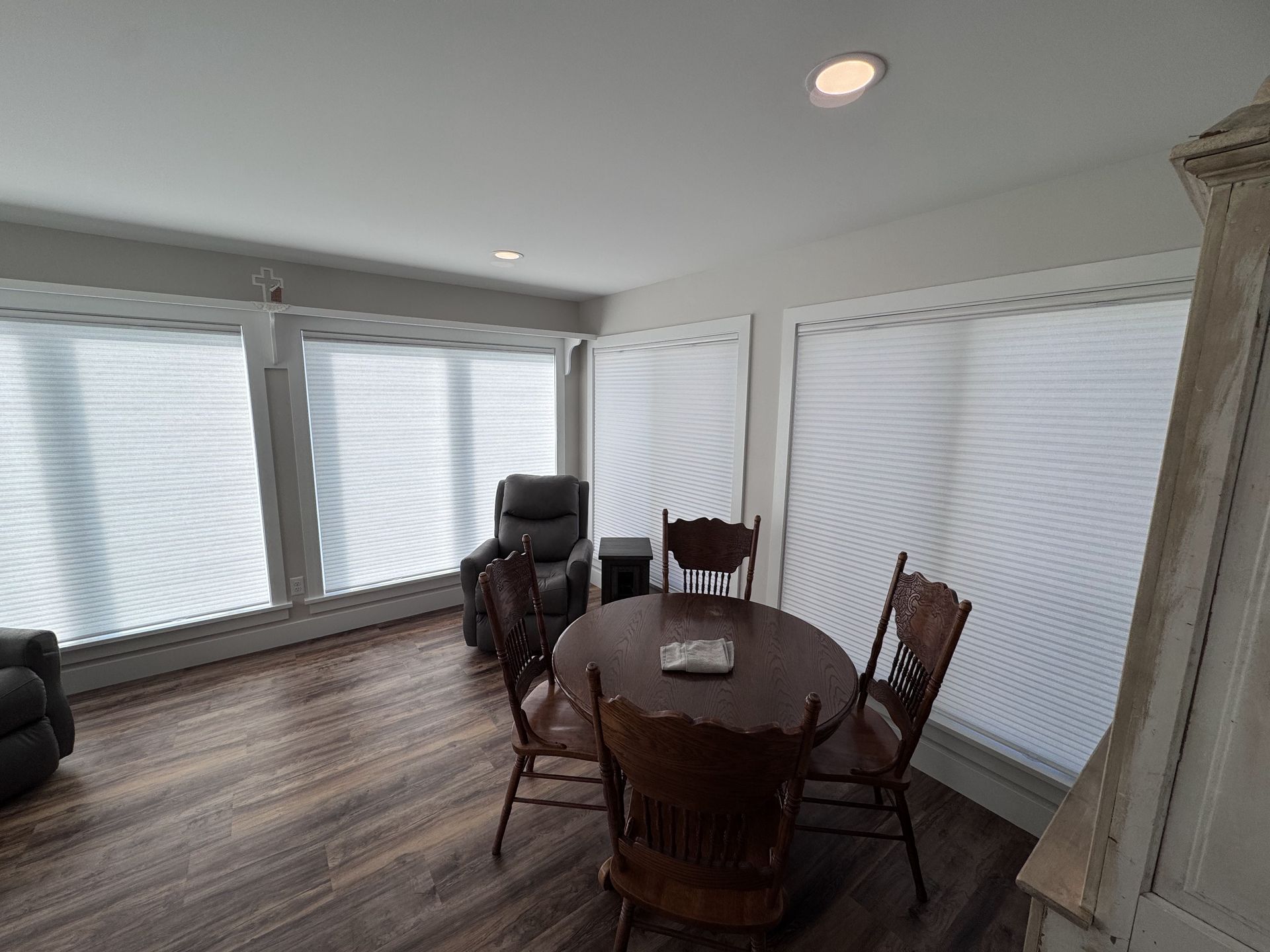 A dining area with a round table, chairs, and window coverings. Brown wood flooring.