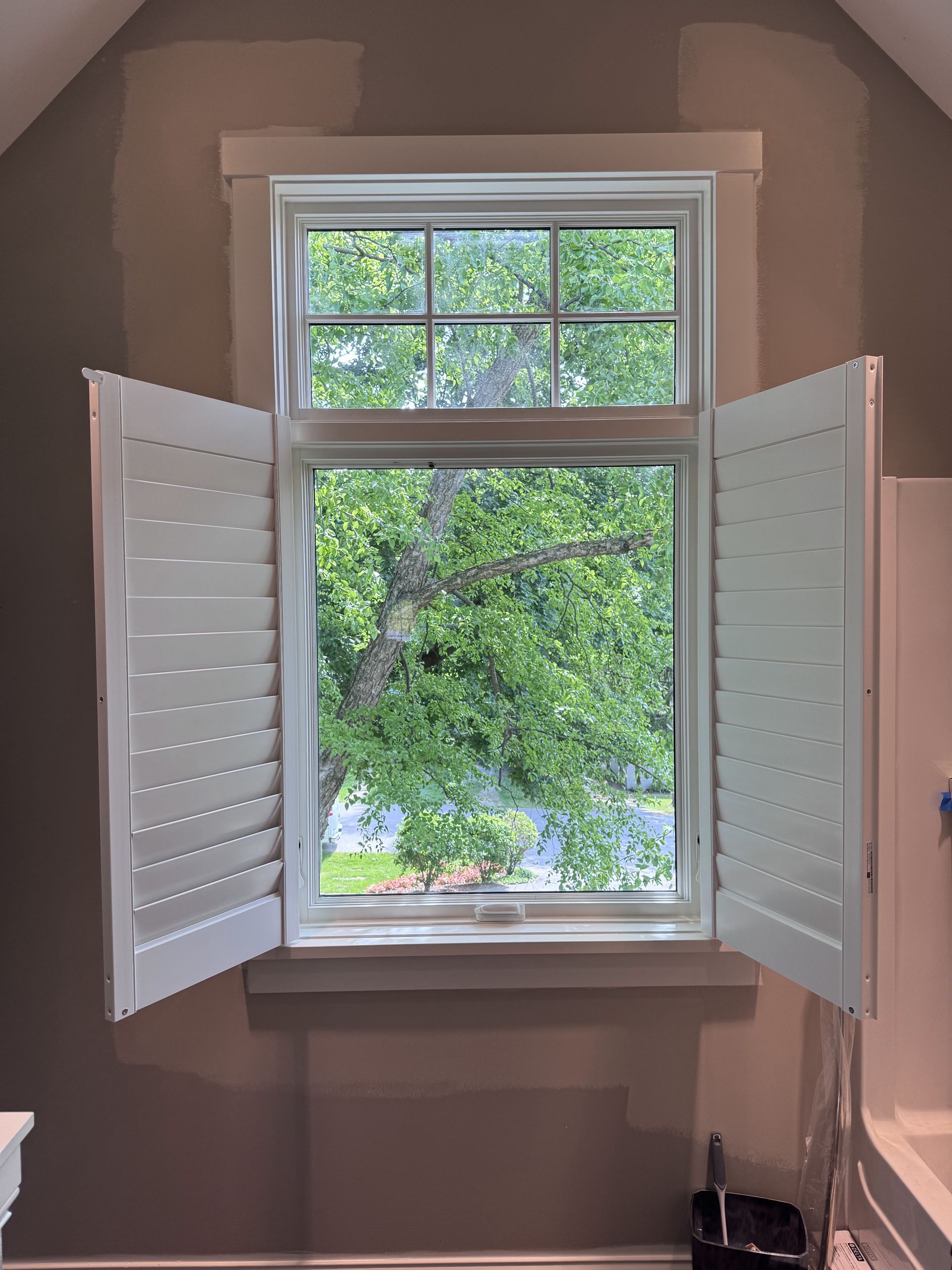 Window with white shutters open, framing a view of green trees, set in a room with gray walls.