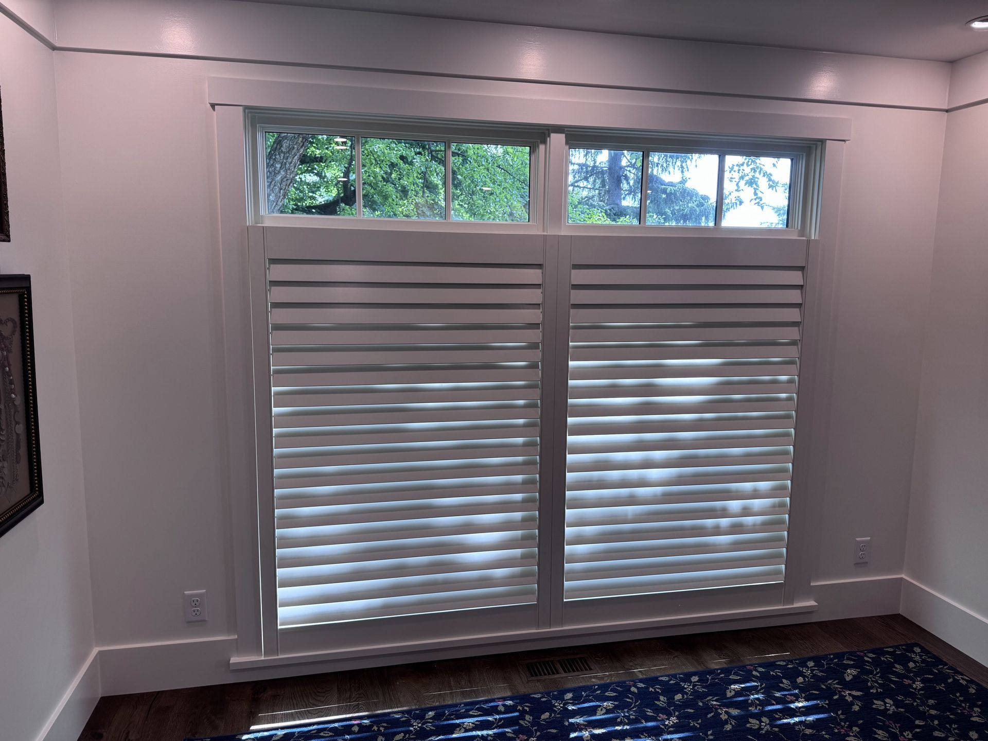 Window with white shutters, revealing a view of green trees.