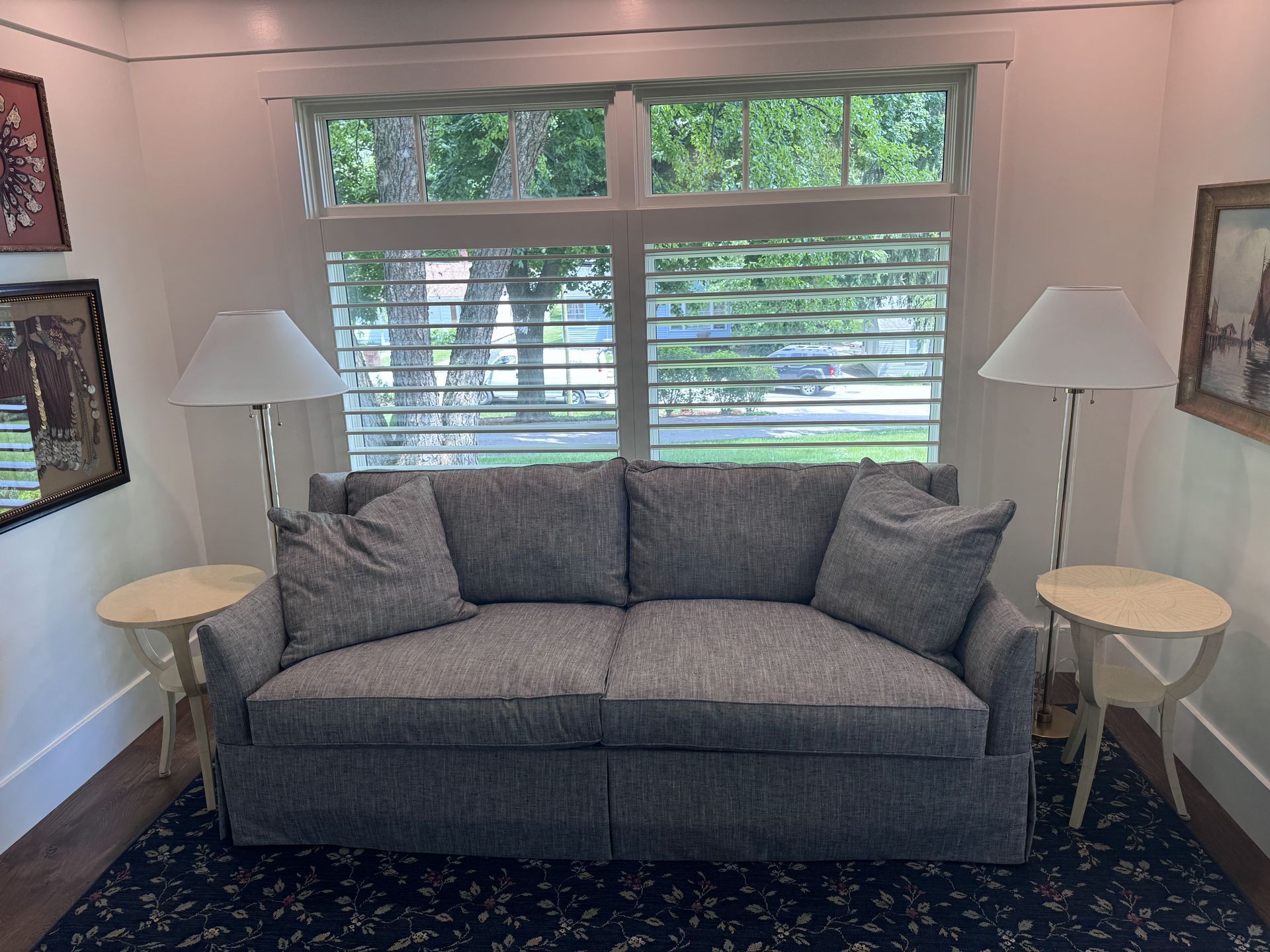Gray patterned sofa with pillows, flanked by lamps and side tables, in front of a window with slatted shutters.