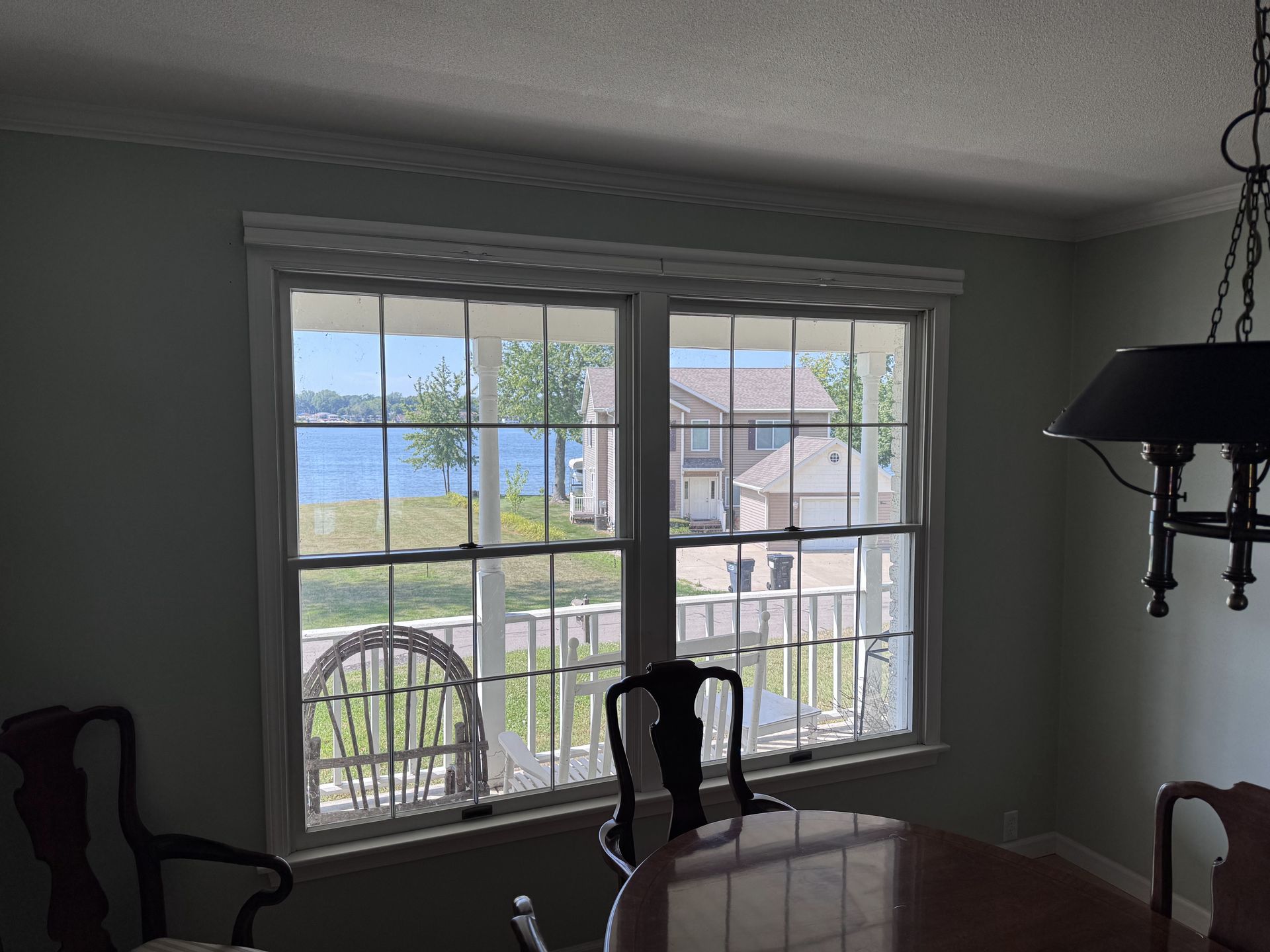 Two-pane window in a dining room overlooking water and houses. Green walls, a table, and chairs are visible.