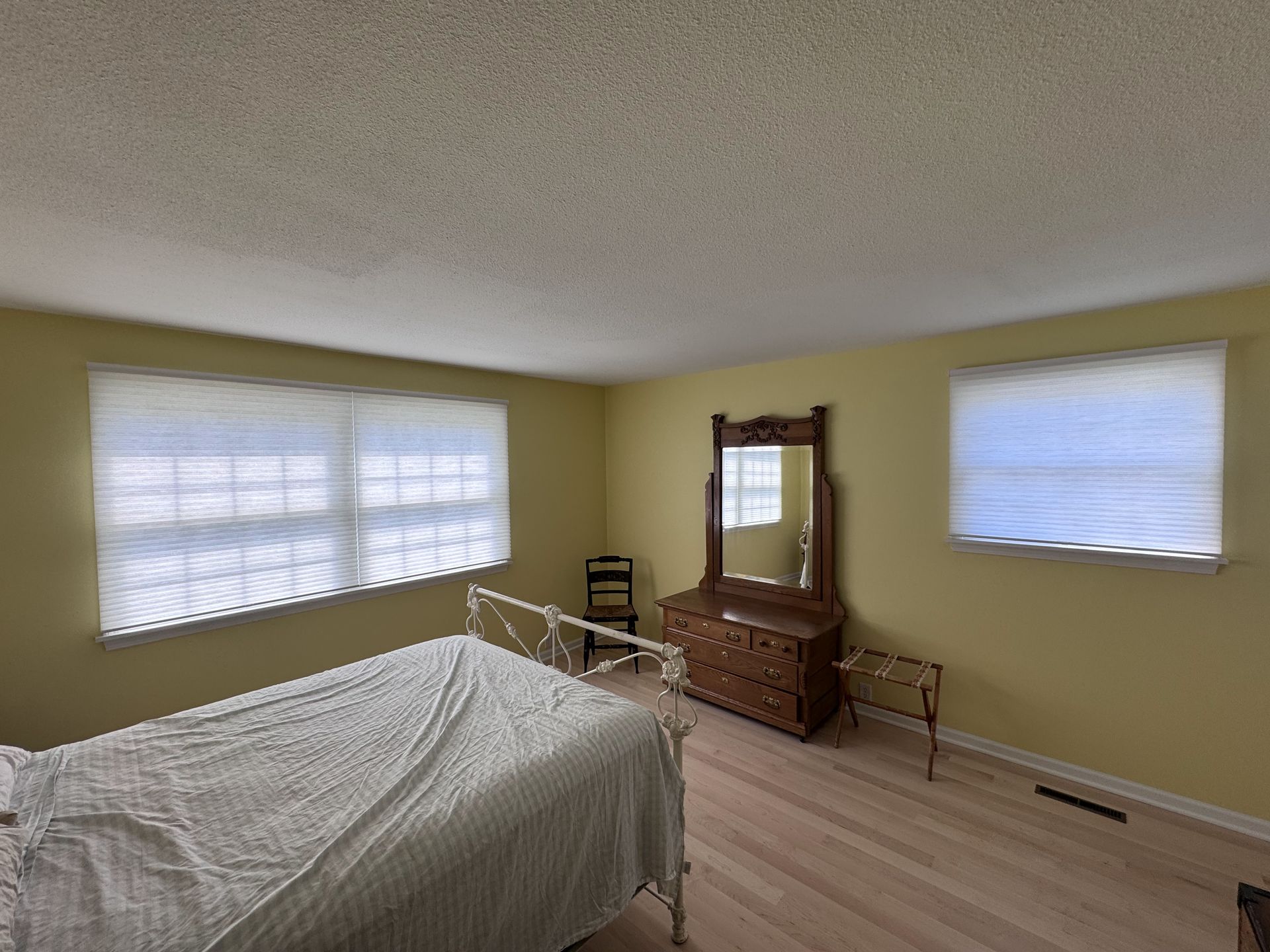 Bedroom with a bed, dresser, and windows. Pale yellow walls, white bedding, and wooden furniture.