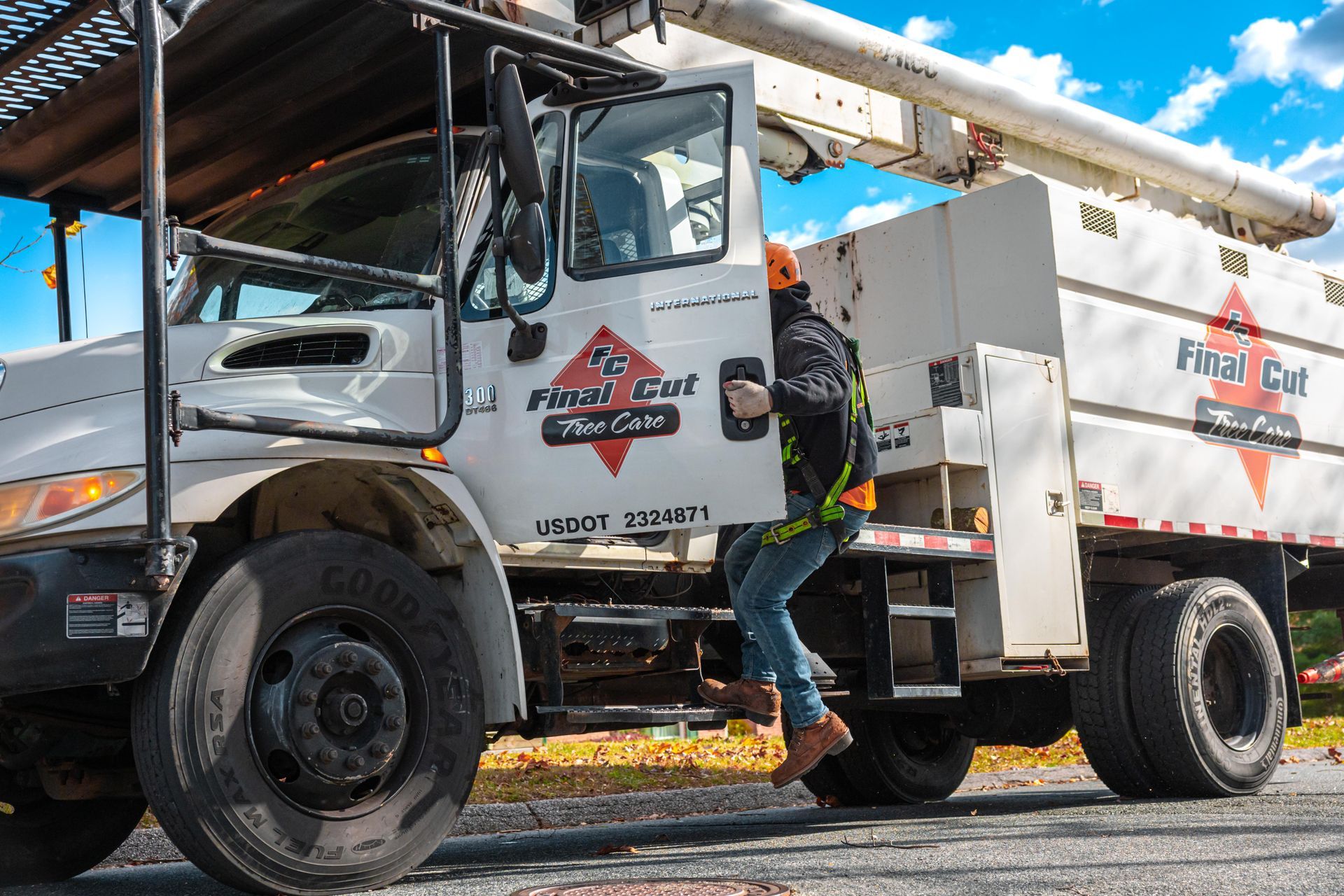 Worker in safety gear entering a white First Cut tree service truck, under a blue sky.
