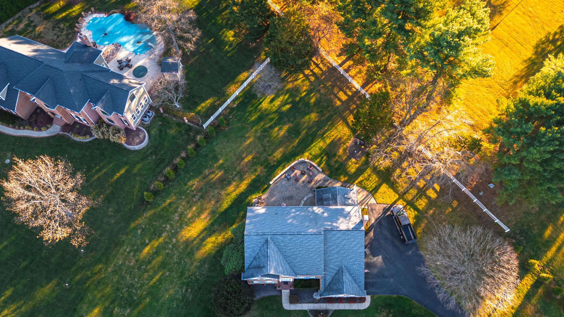 Aerial view of a suburban neighborhood, featuring homes, trees, a pool, and fences bathed in warm sunlight.
