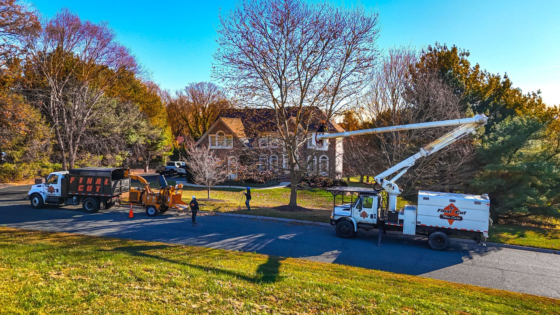 Tree trimming in progress: trucks with lift equipment and a wood chipper in front of a house on a sunny day.