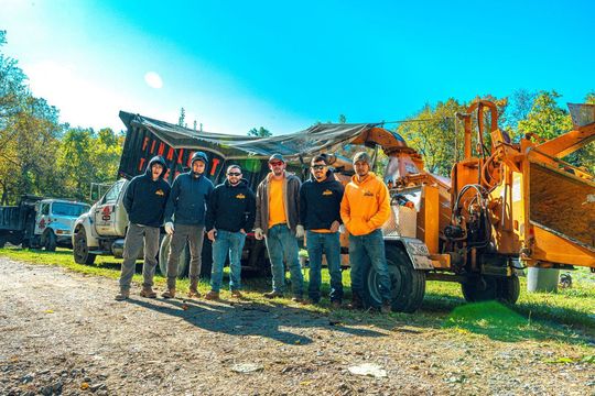 A team of seven in work attire stands by a wood chipper on a sunny day.