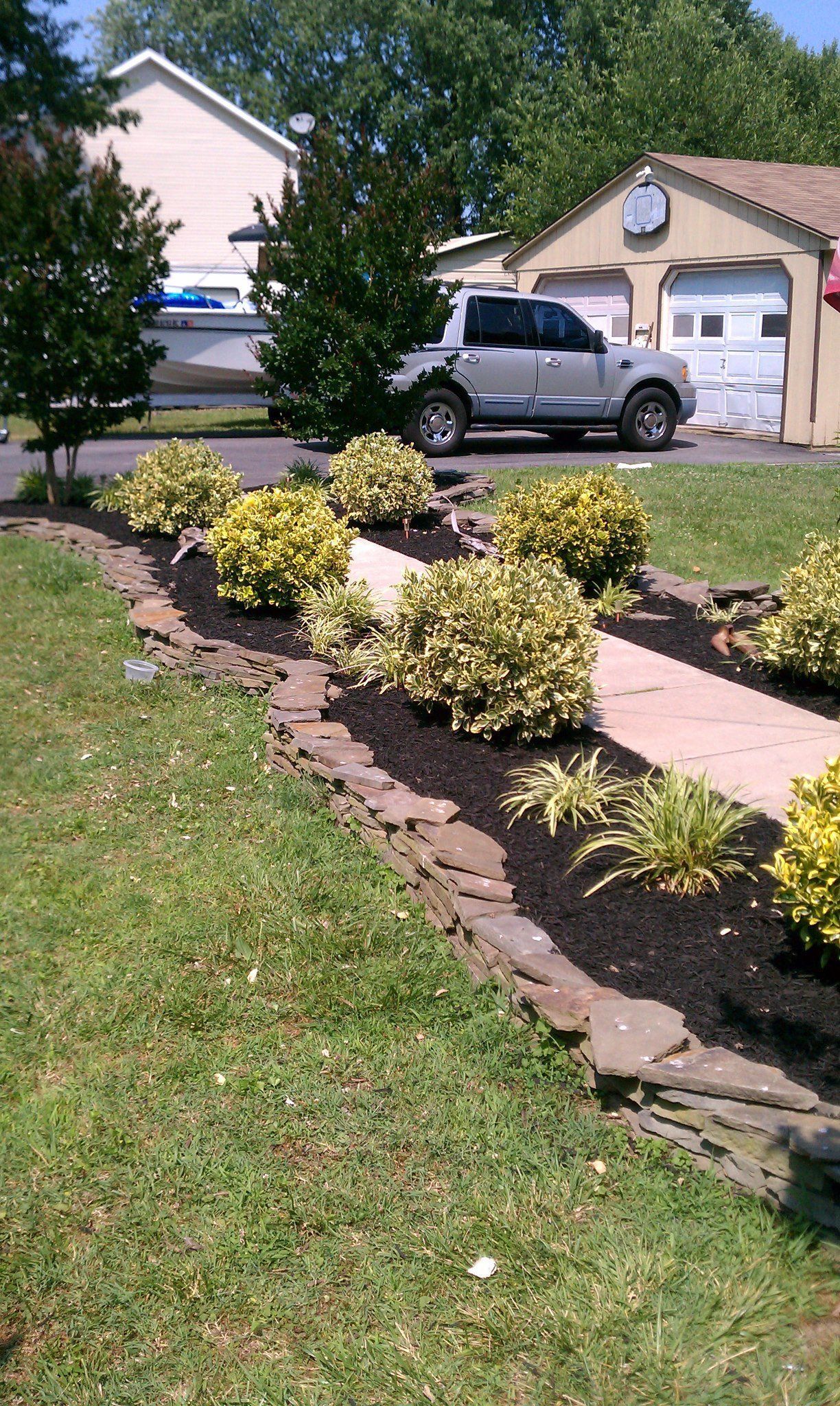 Landscaped yard with stone walkway, black mulch, and yellow-green shrubs. A silver truck is parked on the street.