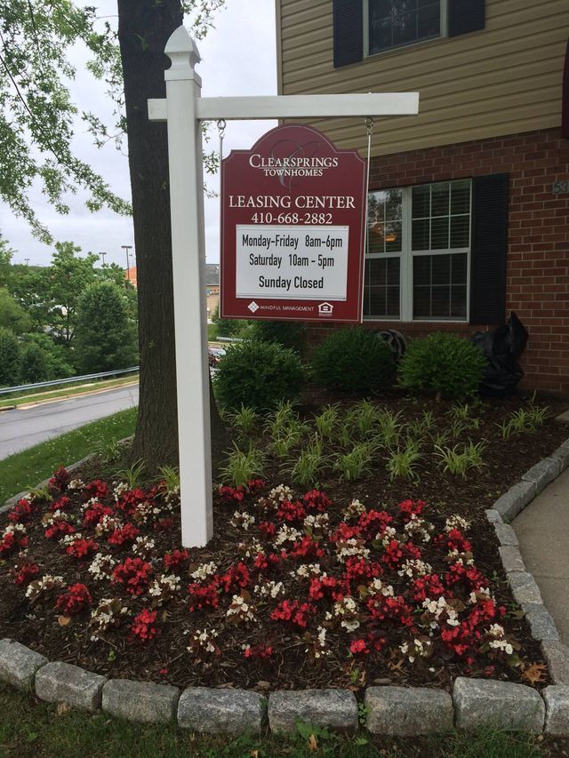 Sign for a leasing center with hours. Sign is red and white, in a flower bed.