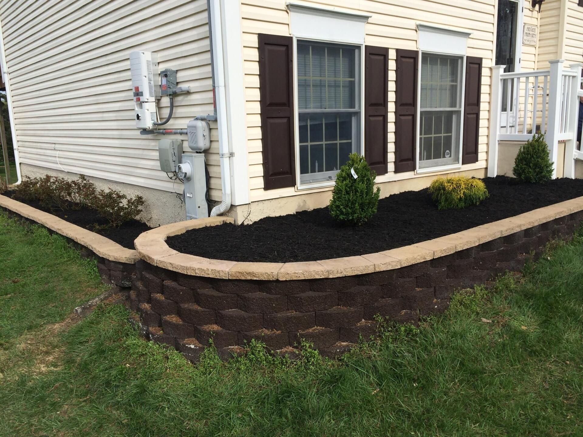 Tan retaining wall with dark mulch and shrubs in front of a beige house.