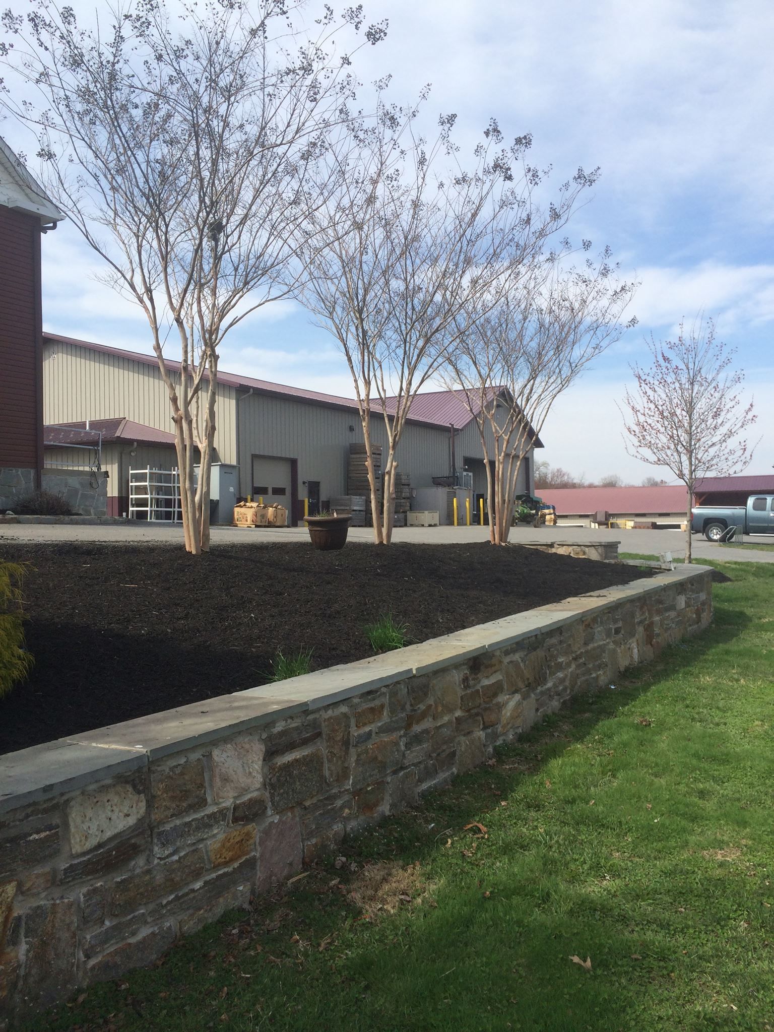 Stone wall with a landscaped area and bare trees in front of a tan building with a red roof.