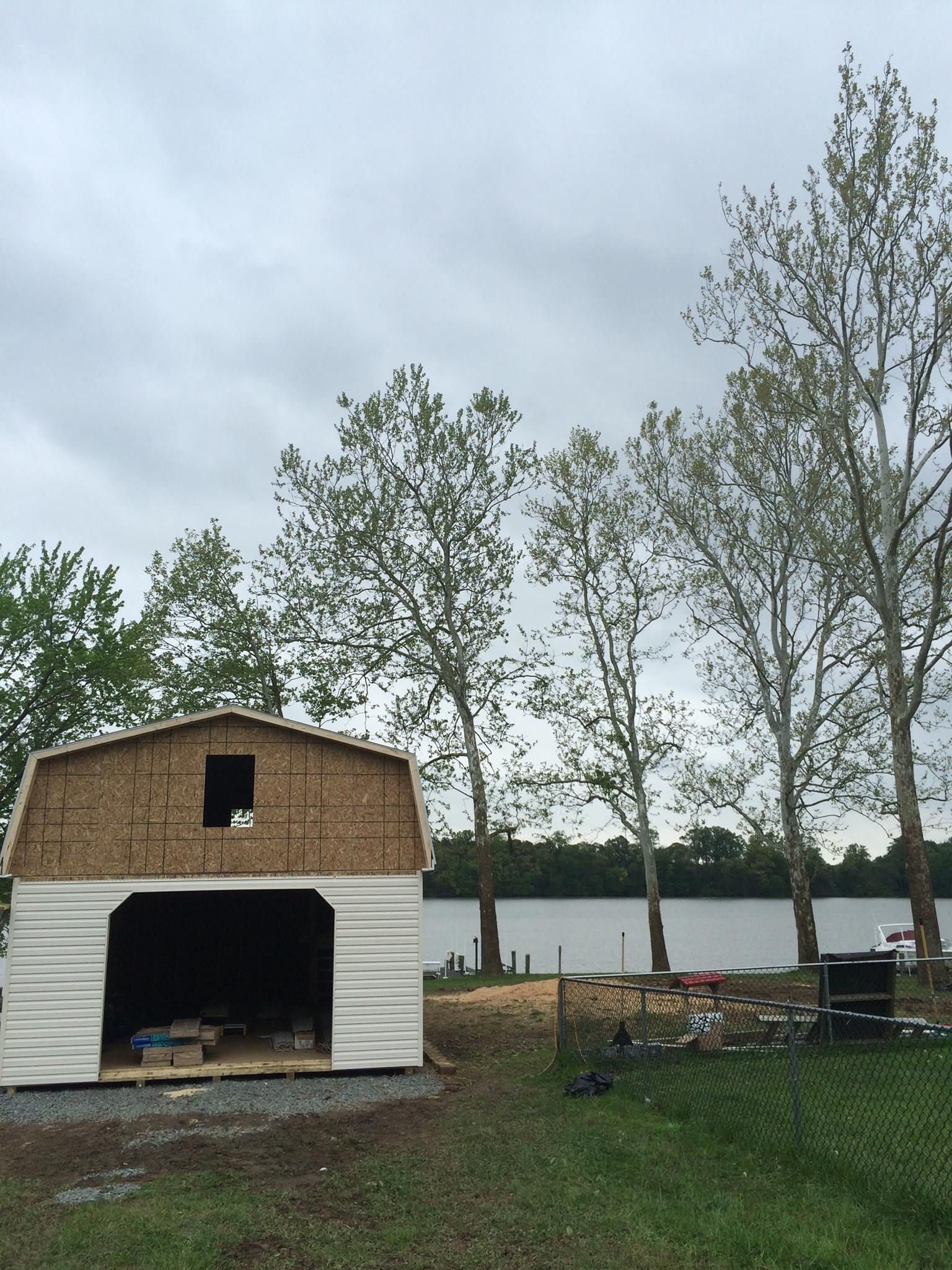Unfinished shed with open front by a lake, under a cloudy sky. Tall trees on the right.