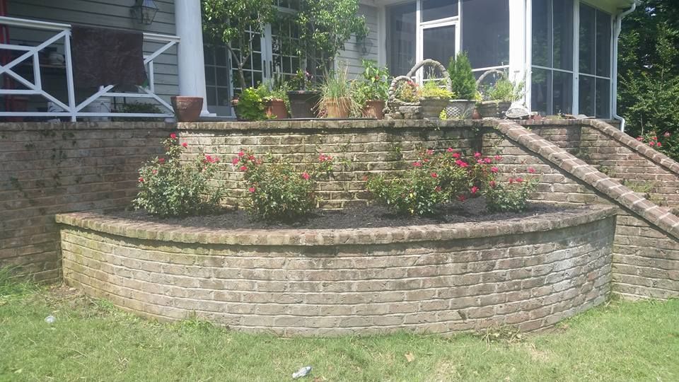 Brick retaining wall with rose bushes and potted plants on a porch.