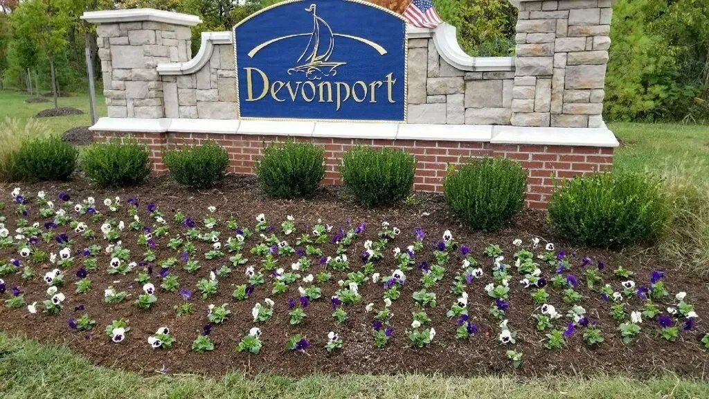 Sign for Devonport with brick base, stone pillars, and flower bed of white and purple pansies.