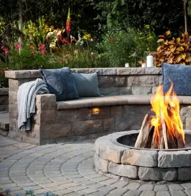 Stone fire pit with burning wood in front of a built-in stone bench with pillows and a blanket in a garden setting.