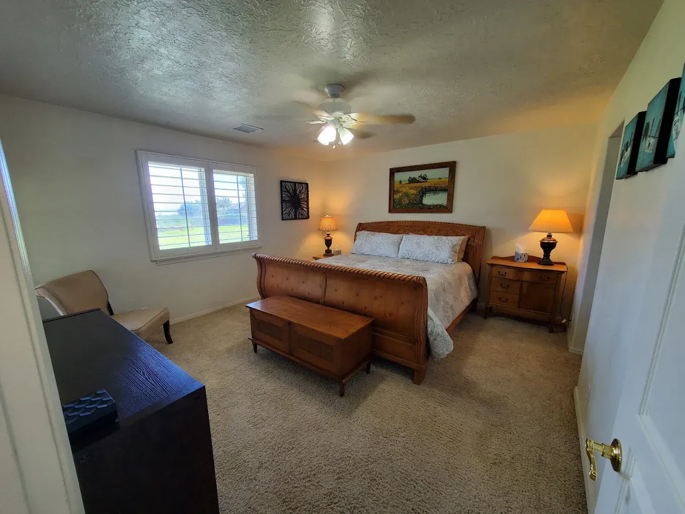 Bedroom with bed, nightstands, chest, and a window. Beige carpet, light walls, lamps.