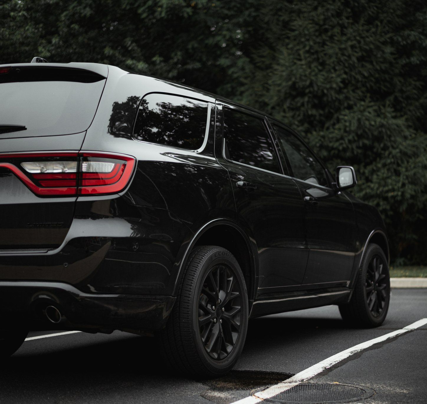 Black SUV parked next to a white line, with dark wheels and a backdrop of green trees.