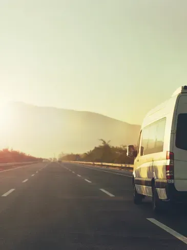 Furgoneta blanca estacionada al costado de una carretera, montaña al fondo, luz del sol.